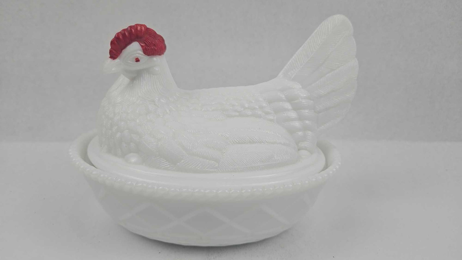 Milk Glass Hen on Nest Covered Dish: This two-piece white milk glass covered dish is modeled in the form of a seated hen resting on a basket-weave nest. The lid is molded with a textured feather motif and features cold-painted red accent