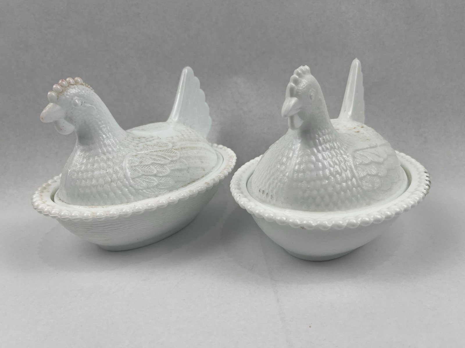 Pair Milk Glass Hen on Nest Dishes: A pair of covered dishes crafted in opaque white milk glass, designed in the traditional hen on nest form. Each object comprises a two-part construction featuring an oval base and a figural lid. The