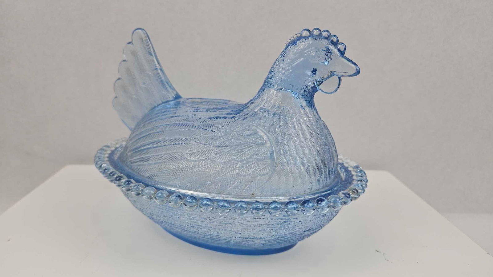 American Blue Pressed Glass Hen on Nest: An American pressed glass covered dish composed of two parts in a translucent light blue color. The figural lid is molded in the shape of a nesting hen, featuring detailed feather textures, a comb, an