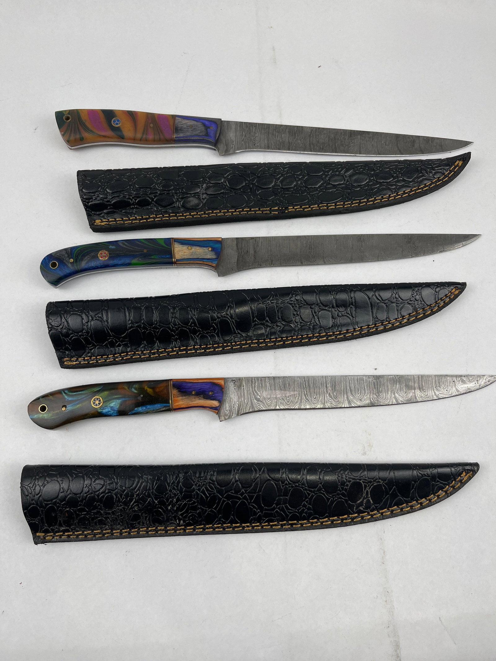 Set 3 Damascus Fillet Knives w/ Sheaths: A trio of custom fixed-blade knives, designed for filleting or slicing, each accompanied by a matching sheath. The blades are constructed of pattern-welded Damascus steel, exhibiting a high-contrast w