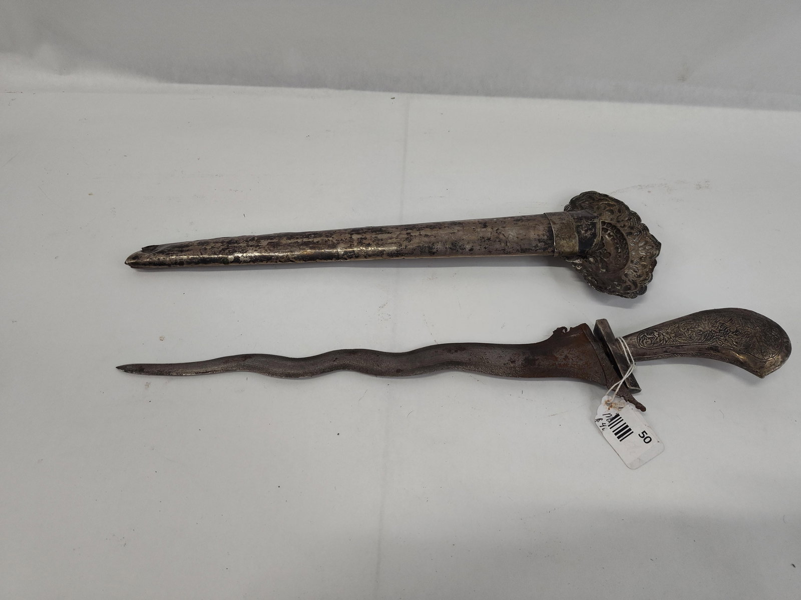 Indonesian Kris w/ Repousse Sheath: This kris features a steel blade of luk or wavy form, characterized by visible forging patterns and a slight taper toward the point. The blade is fitted to a hilt of non-ferrous, silver-colored
