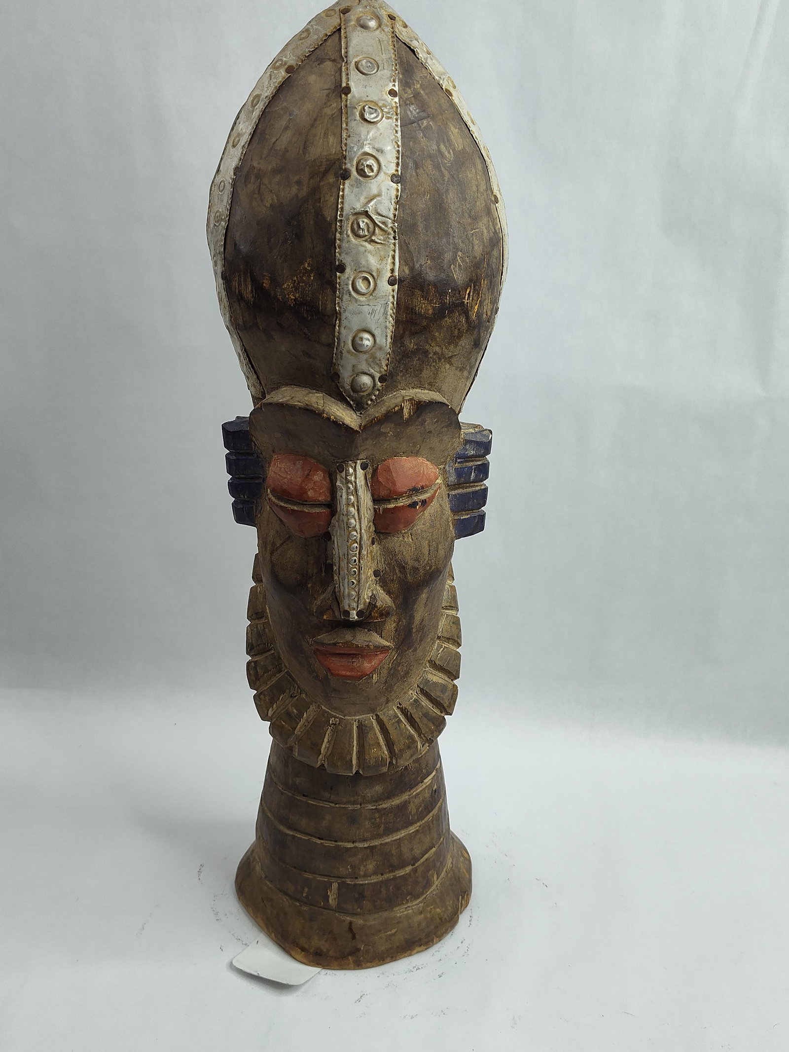 Carved Wood & Metal Figural Sculpture: A tall, hand-carved wooden figural sculpture featuring a vertical, elongated form with stylized anthropomorphic features. The head is topped with an oversized, miter-like headdress or coiffure, which 