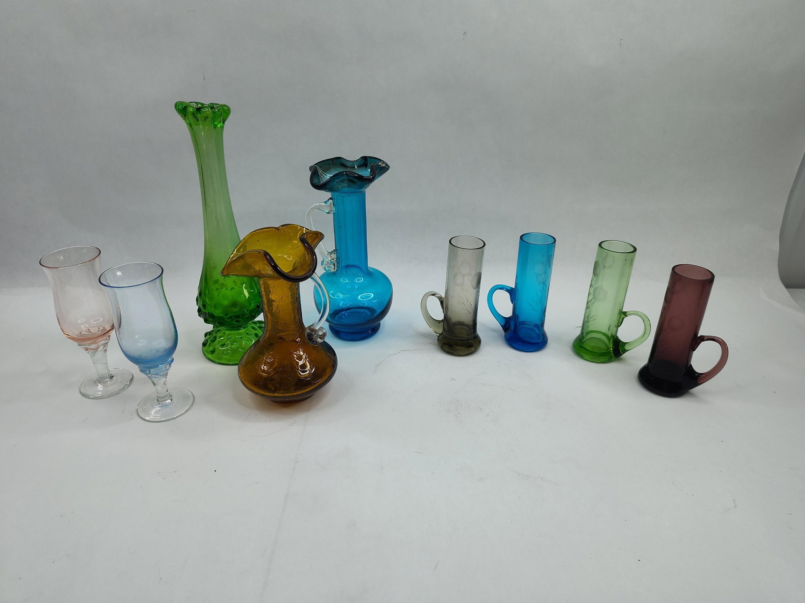 Set Mid-C. Glass Vessels: A collection of various mid-century and decorative glass vessels in a range of colors and forms. The assortment includes a tall, green swung vase featuring a hobnail patterned base and a ruffled rim. 