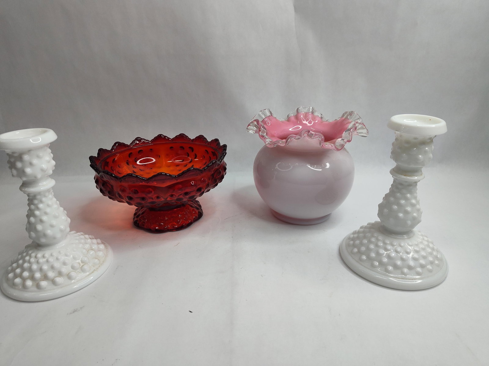Fenton Glass Hobnail Tableware Set: A collection of four American art glass pieces produced in the hobnail pattern. The group includes a pair of milk glass candlesticks, a red-to-amberina pedestal bowl, and a cased glass ruffled vase.