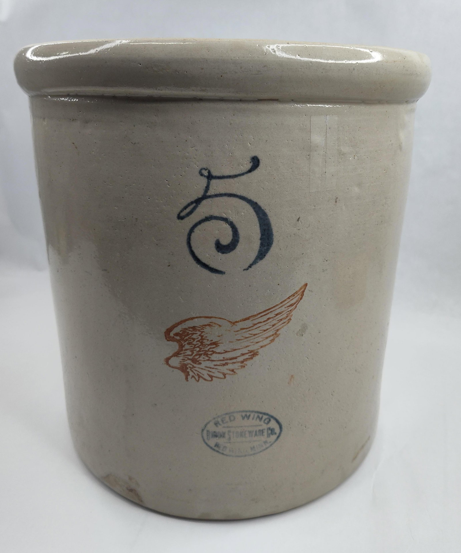 Red Wing Union Stoneware 5 Gal. Crock: This five-gallon American stoneware crock features a cylindrical form composed of salt-glazed or light grey slip-finished clay. The exterior is decorated with three distinct cobalt and iron-oxide elem