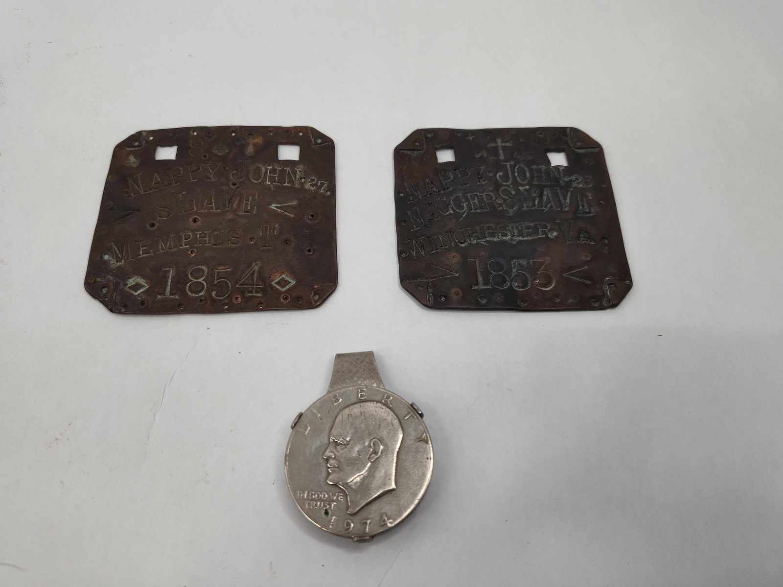 2 Copper Alloy Tags & 1974 Dollar Clip: A collection of three metal objects consisting of two hand-stamped copper-alloy rectangular tags and one circular coin pendant. The first copper-alloy tag features hand-stamped characters including