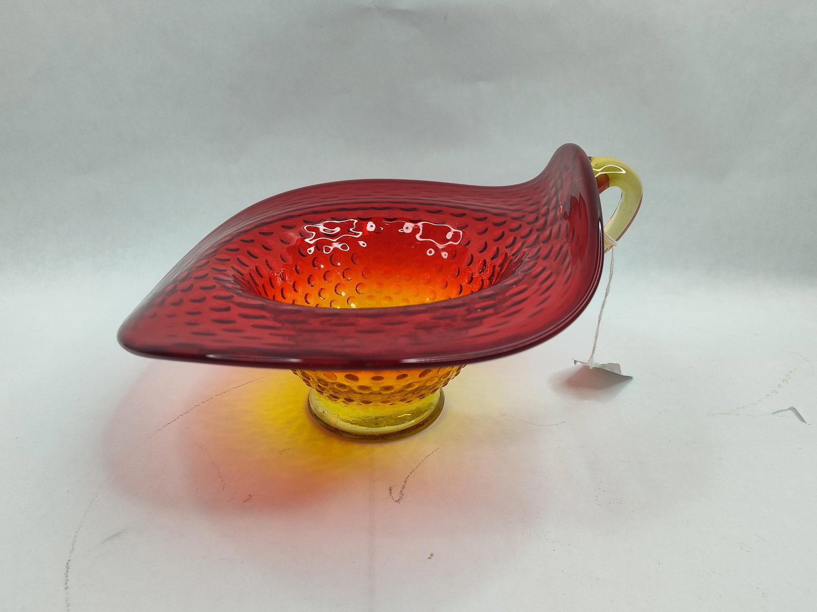 Amberina Glass Hobnail Handled Nappe: A decorative glass nappe or bowl featuring a vibrant amberina colorway. The piece transitions from a translucent yellow at the circular pedestal base to a deep, saturated red at the flared rim. The