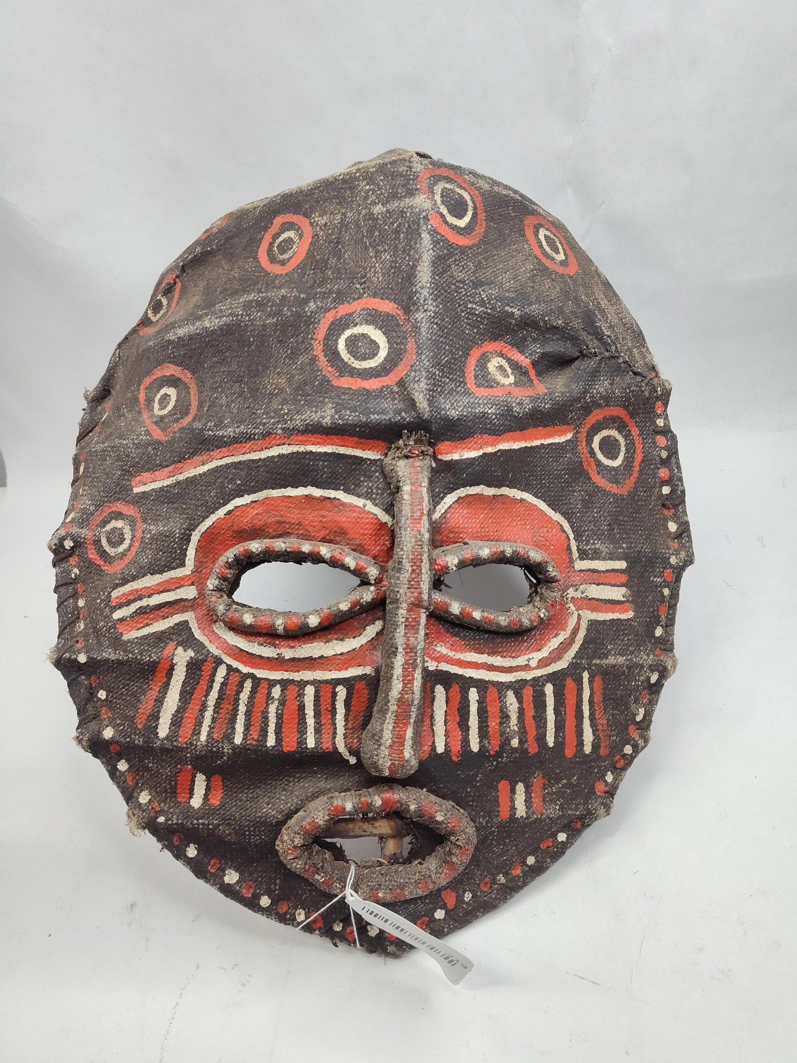 African Bark Cloth Mask w/ Vine Frame: This ethnographic mask is constructed from a dark brown, hand-woven fiber fabric, likely bark cloth, stretched over a structural armature of bent wood and vine. The face features a prominent vertical