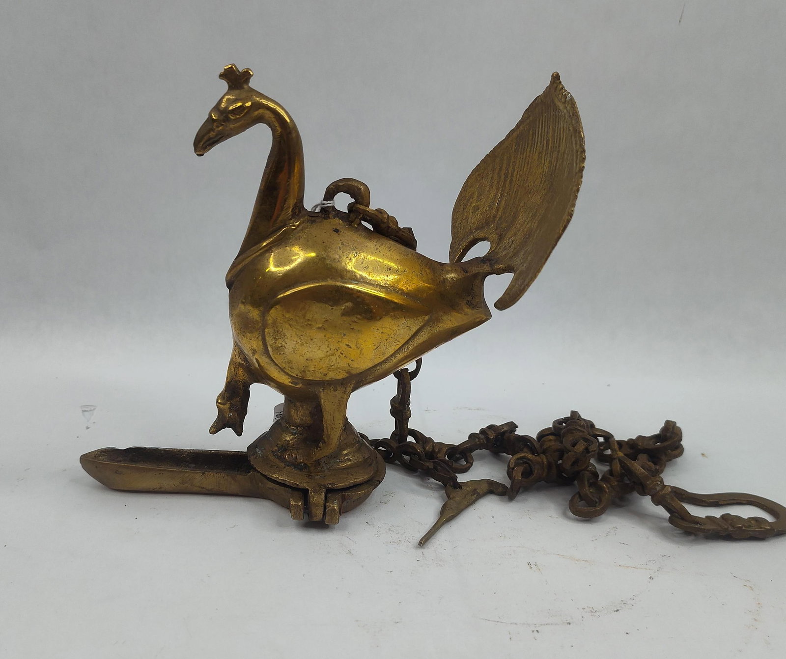 Cast Brass Peacock Hanging Oil Lamp: A cast brass hanging oil lamp, or mayur diya, modeled in the form of a peacock. The piece features a hollow bird-shaped reservoir and an elongated, hinged wick tray at the base. The peacock is cast