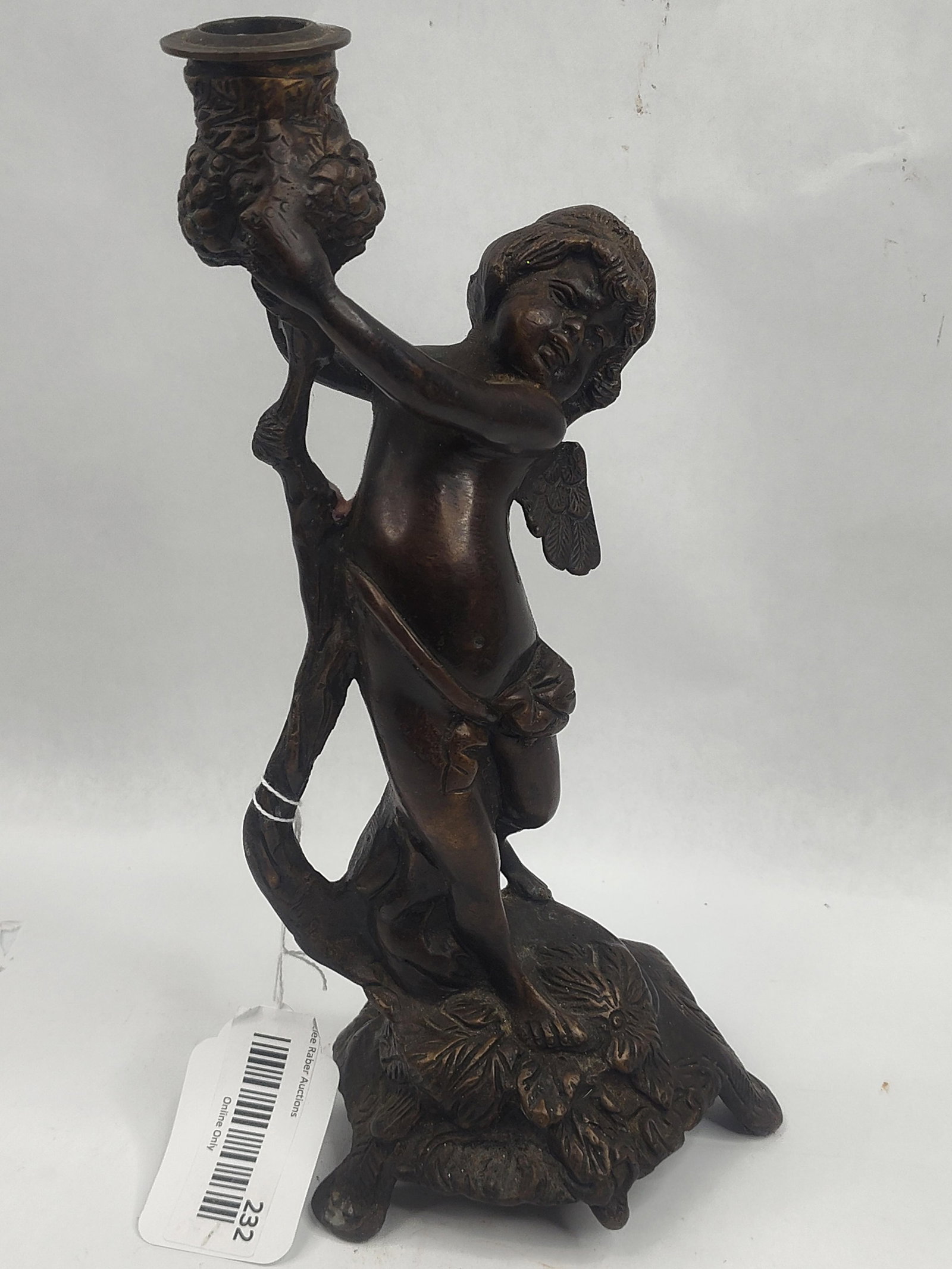 Bronze Figural Putto Candlestick: A patinated cast bronze figural candlestick modeled in the Rococo style. The piece features a winged putto or cherub positioned dynamically against a gnarled tree trunk. The figure supports an urn-for