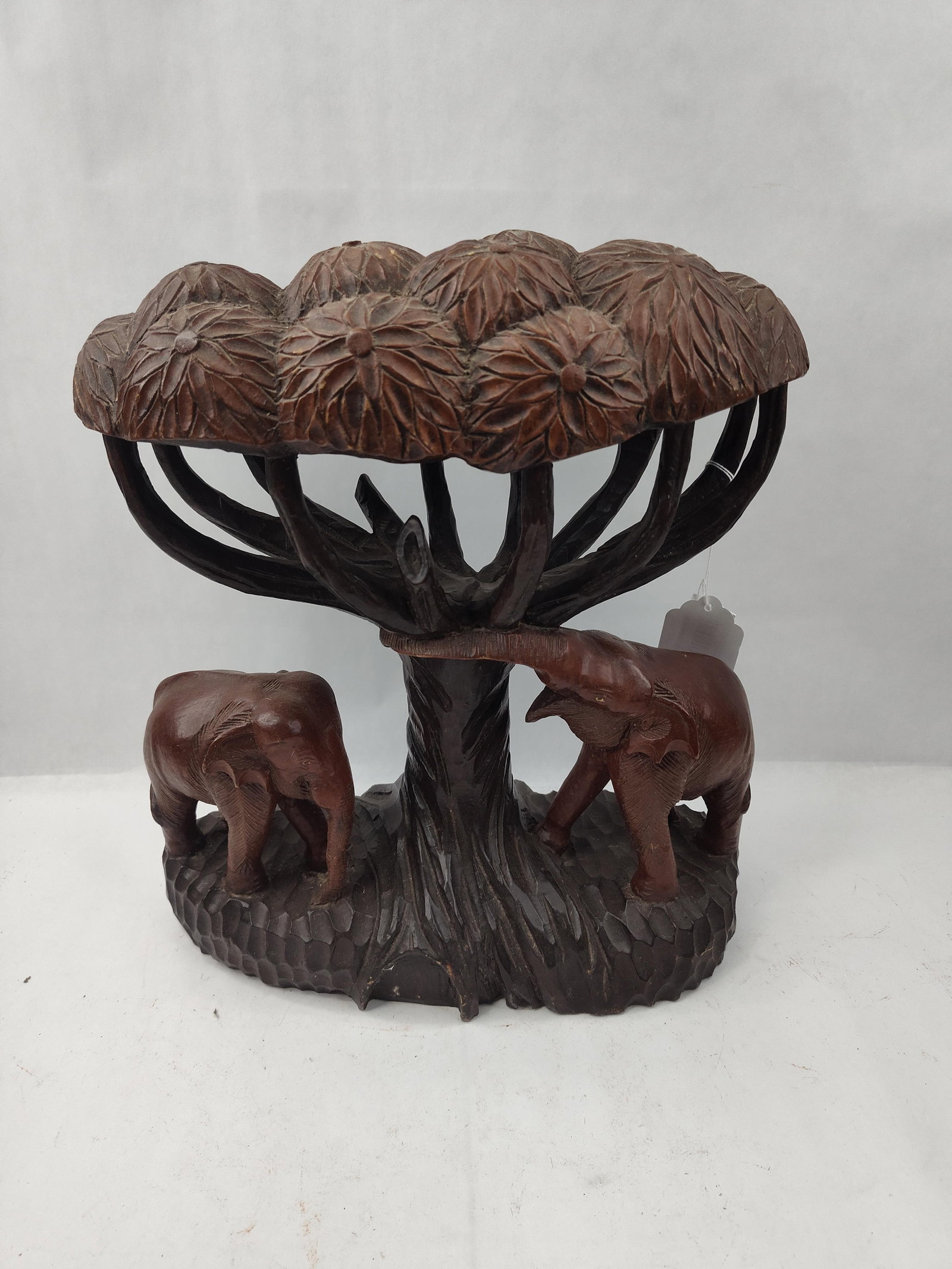 Carved Wood Sculpture of Elephants: A hand-carved dark reddish-brown wood sculpture featuring two elephants standing beneath a stylized tree canopy. The central tree trunk divides into multiple upward-curving branches that support a
