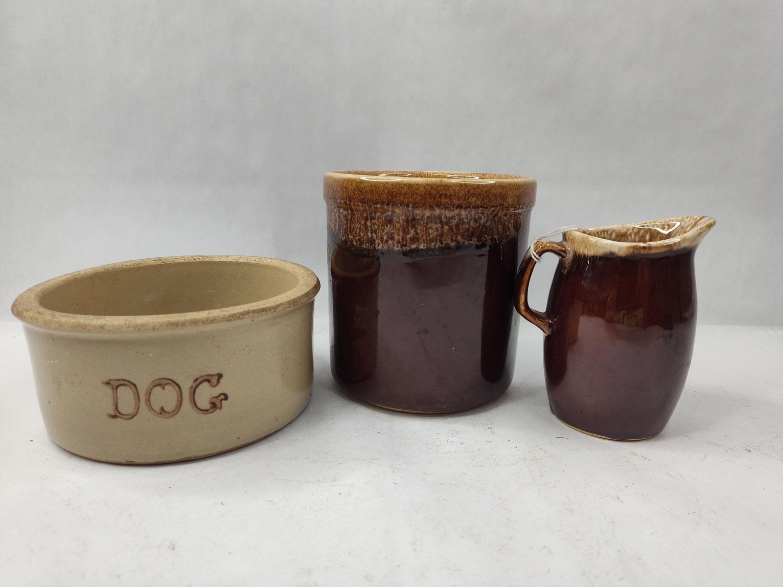 Trio American Brown Glazed Stoneware: This group comprises three pieces of functional American stoneware, each featuring a variation of a brown slip glaze. The first item is a shallow, oval-formed tan stoneware pet feeder. The exterior is