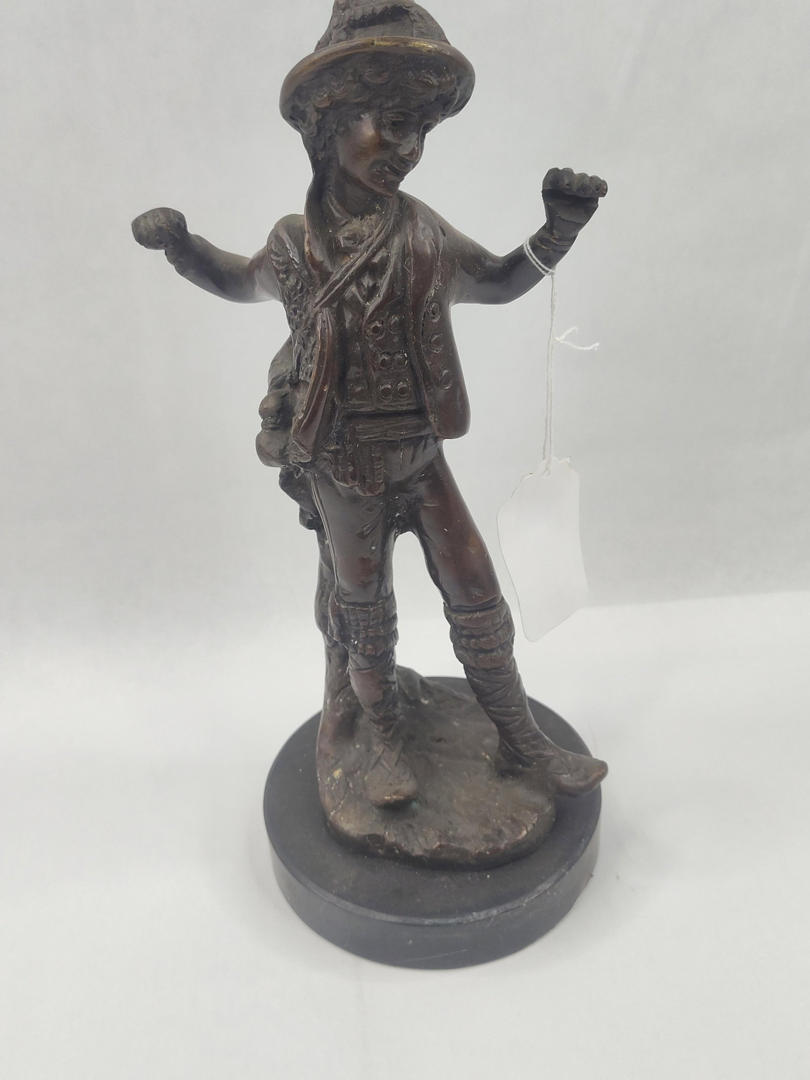 Cast Metal Figure of a Dancing Boy: This cast metal statuette with a dark brown bronze-toned finish depicts a young boy in rustic European attire. The figure is modeled in a dynamic, celebratory pose with both arms raised and fists