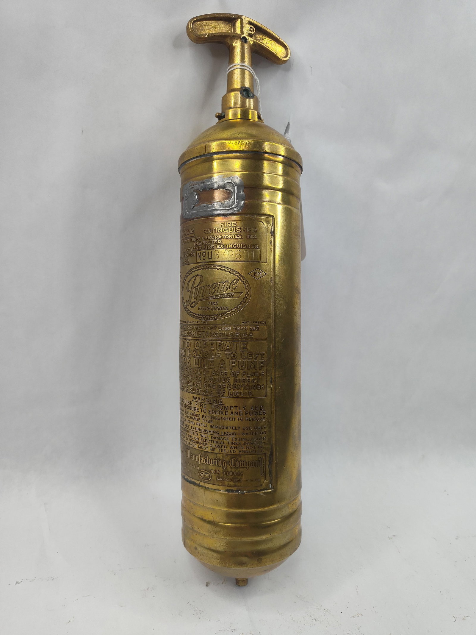 Pyrene Brass Hand Pump Extinguisher: This 1-quart hand pump fire extinguisher is constructed from heavy-gauge brass with a cast metal T-handle plunger at the top. The cylinder body is adorned with a large, rectangular embossed brass