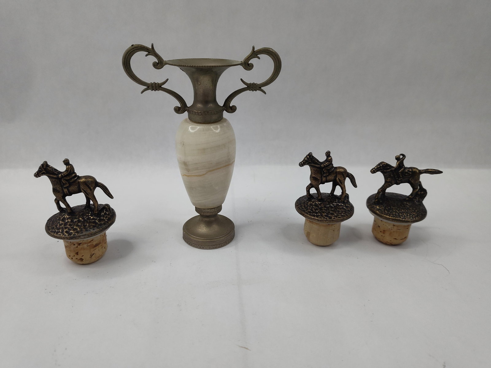 Equestrian Barware & Neoclassical Urn: A collection of barware and decorative items comprising three bottle stoppers and a twin-handled trophy-form vase. The stoppers feature cast metal figurines of jockeys on horseback, each captured in