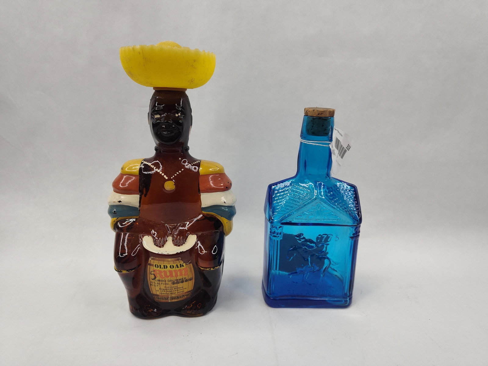 Pair Glass Bottles: Rum & Syrup: A group of two glass bottles of different forms and colors. The first is an amber glass figural bottle depicting a man in colorful attire, functioning as a drummer with hands resting on a barrel. It