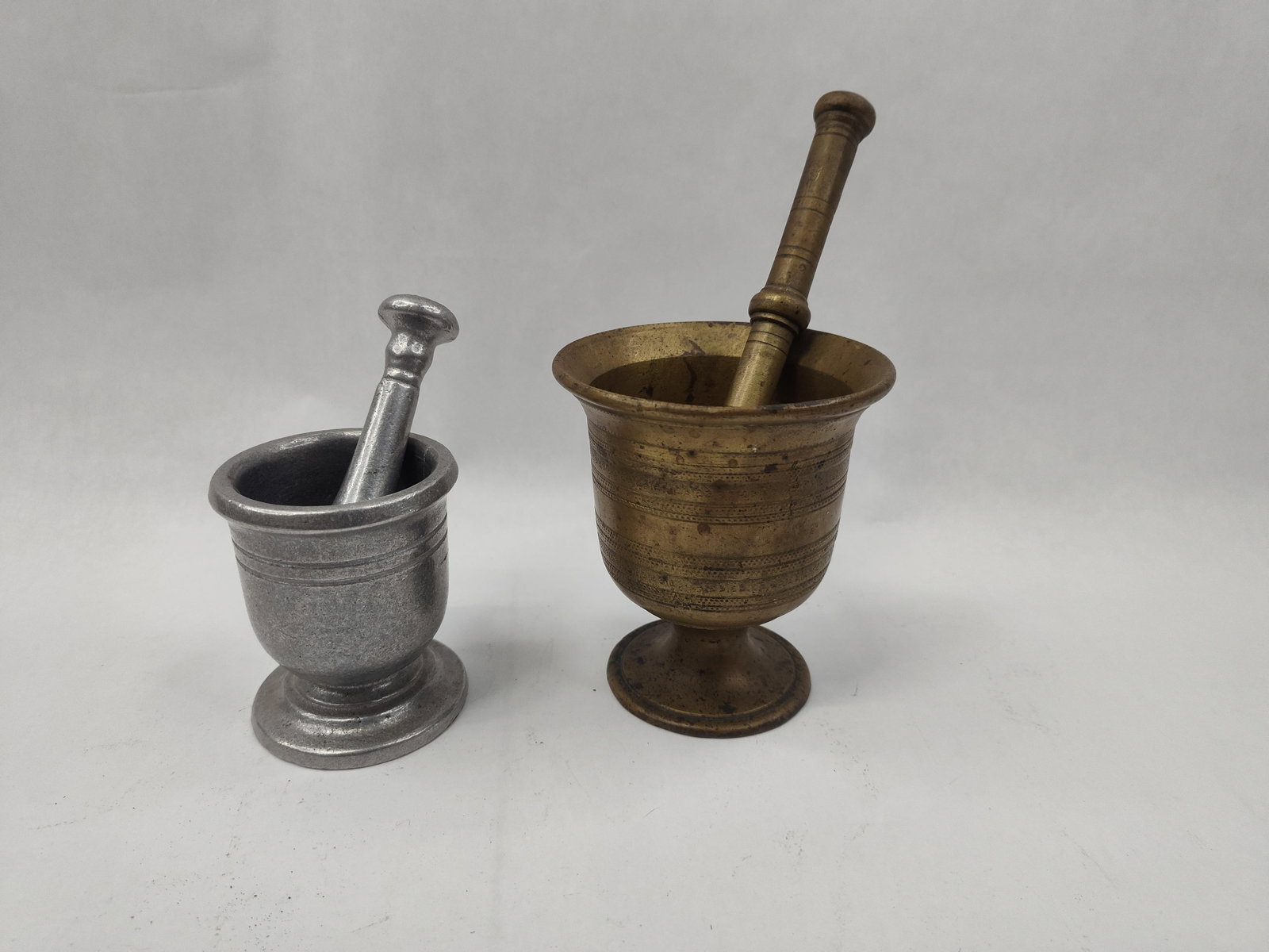 Pair American Metal Mortar & Pestle: A group of two mortars and pestles of differing metals and sizes. The larger set is constructed from a yellow-toned cast metal, likely brass or bronze, featuring a footed mortar decorated with