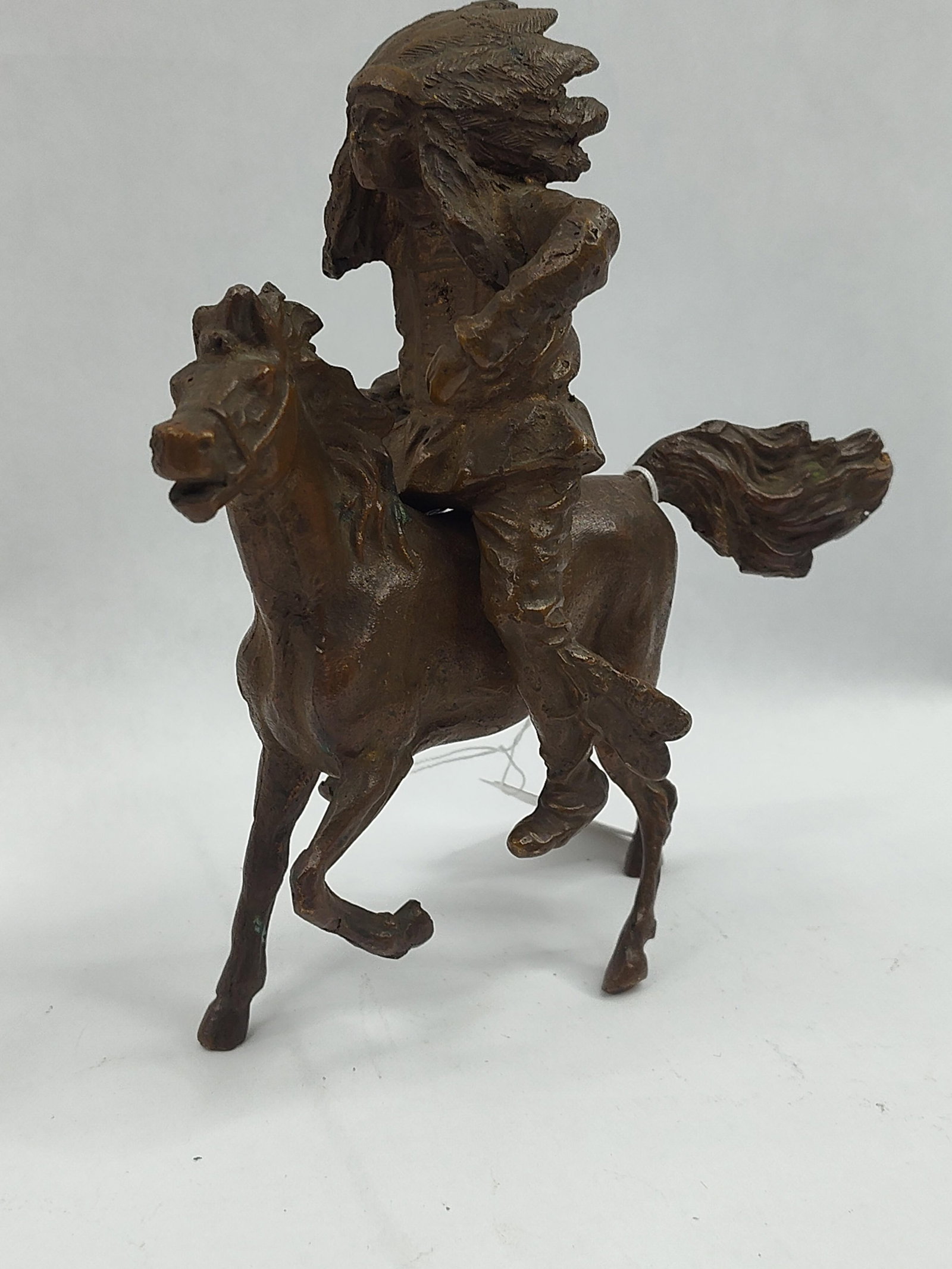 Native American Warrior Cast Sculpture: A cast metal sculpture, likely bronze or a copper alloy, depicting a Native American figure in a traditional feathered headdress mounted on a galloping horse. The work is executed in a rustic, highly