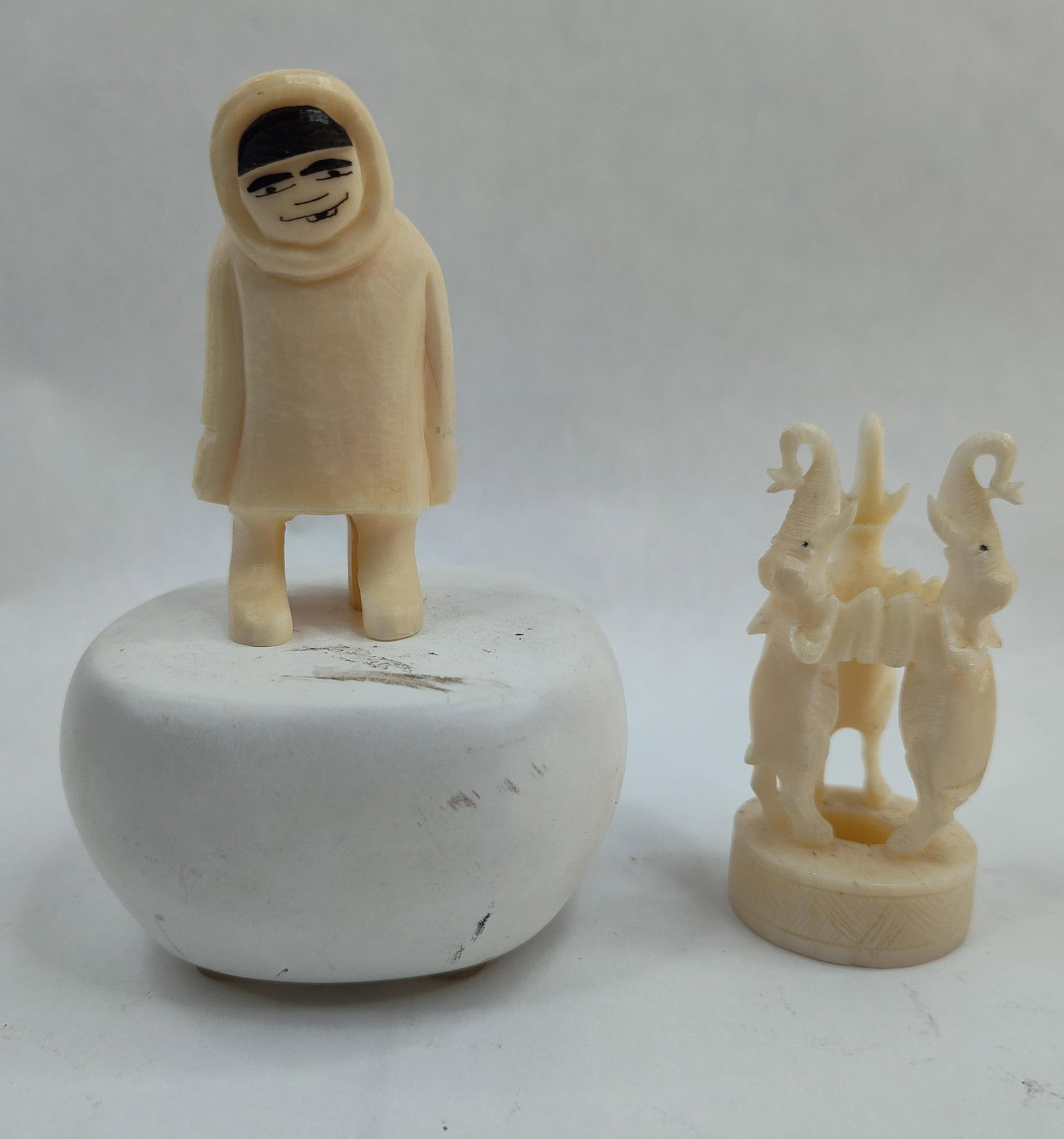 Two Carved Norwegian Bone Figurines: A Norwegian carved bone or ivory-toned synthetic sculpture of a stylized human figure wearing a hooded parka, accompanied by a smaller carved figurine of three elephants. The larger figure features