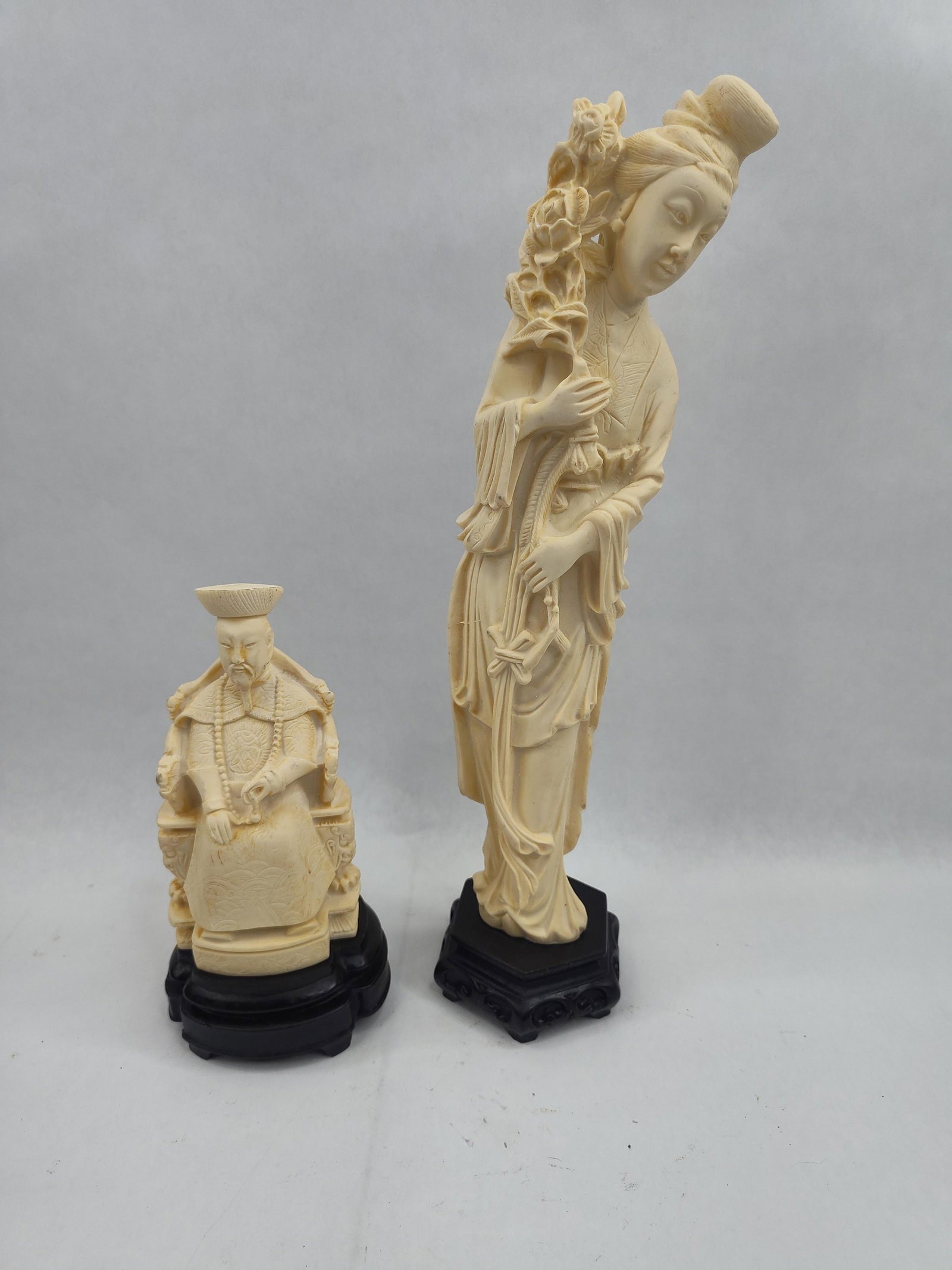 Pair Chinese Figural Faux Ivory Resins: A group consisting of two Chinese-style figural sculptures crafted from a cream-colored cast resin material designed to simulate carved ivory. The first figure depicts a seated male dignitary or