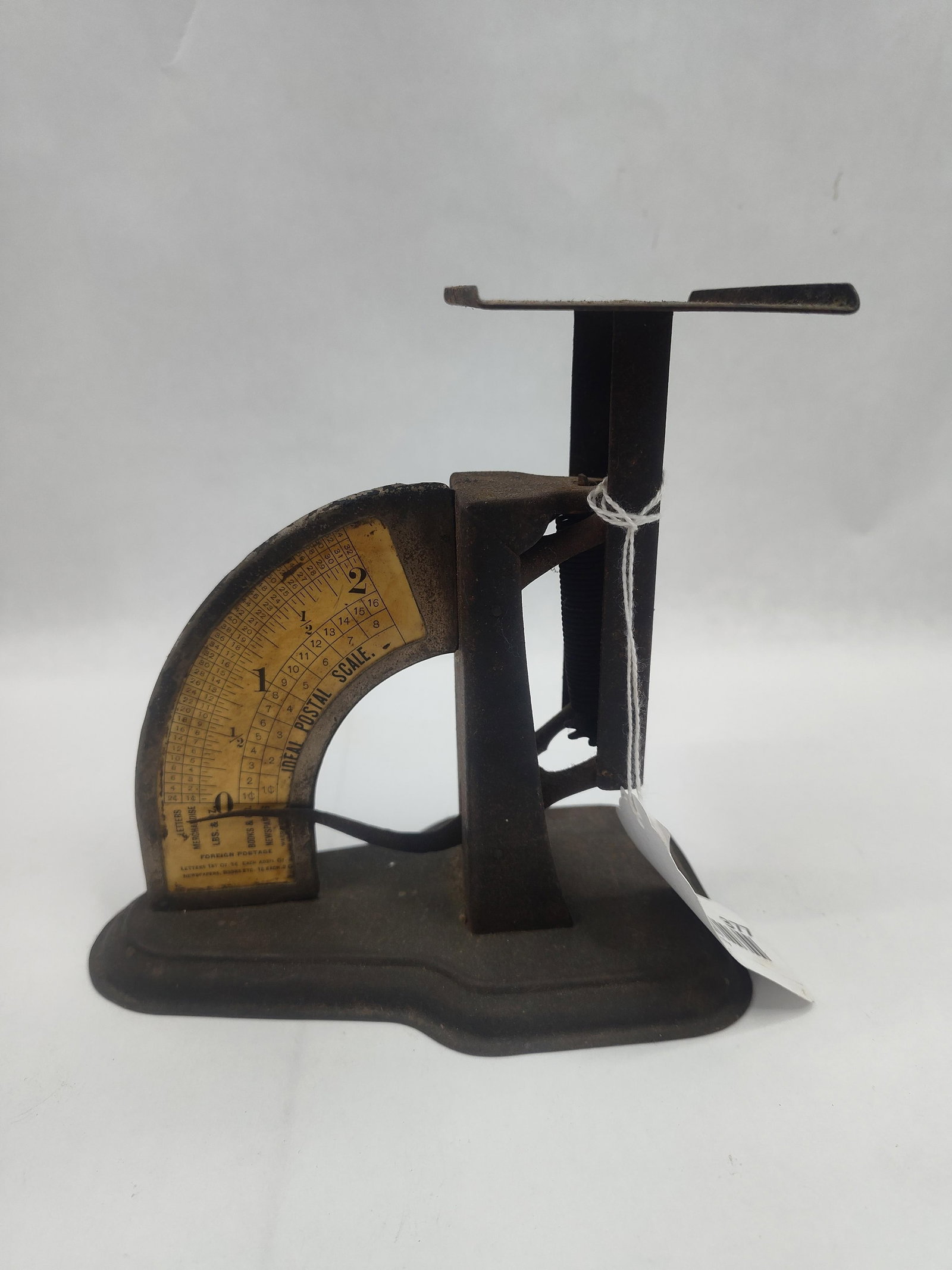 Ideal Postal Scale: A vertical pendulum-style desk scale of metal construction, featuring a dark aged finish. The scale utilizes a flat rectangular top platform supported by a vertical member that drives a curved metal