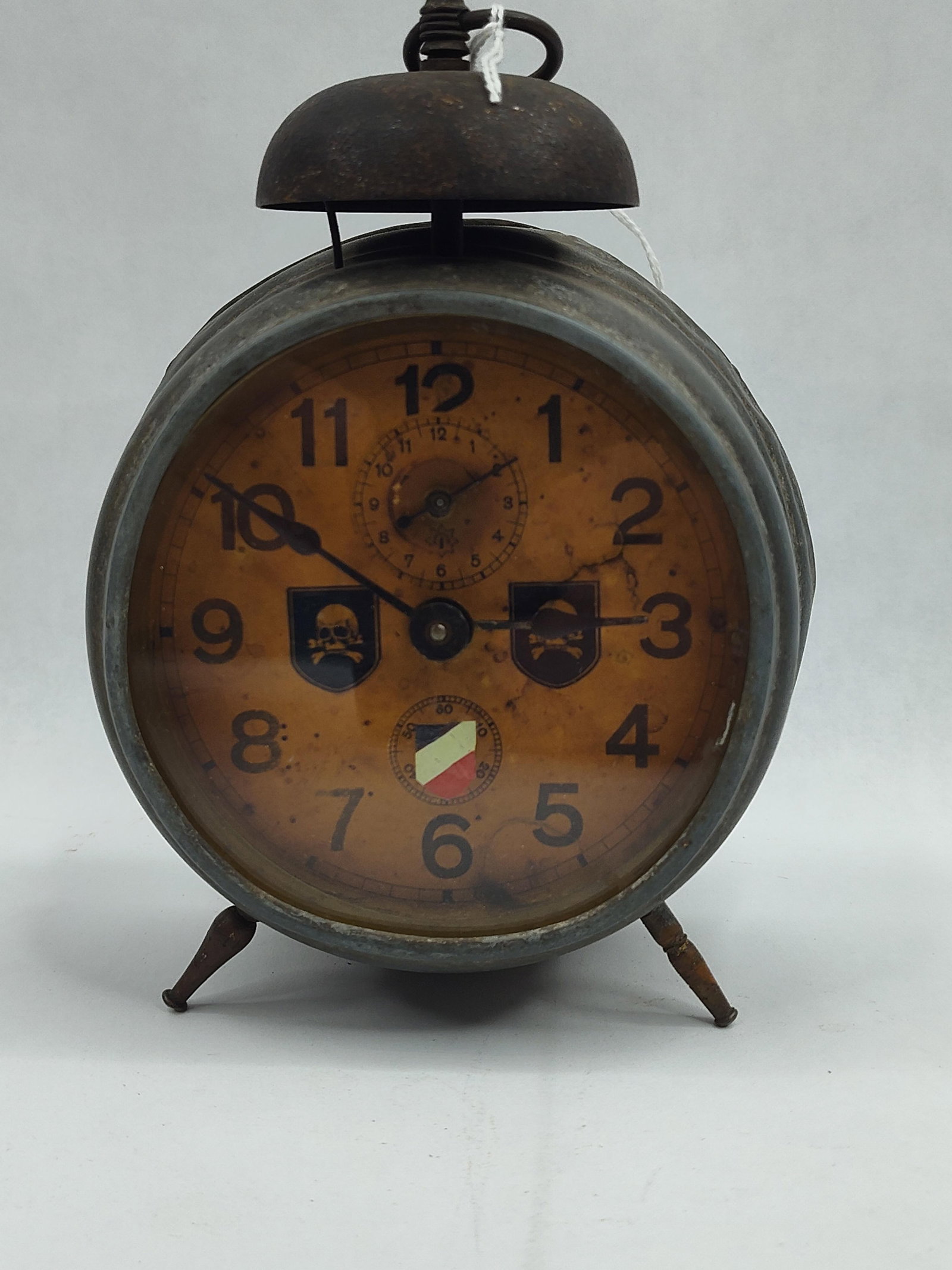 German Mechanical Skull Alarm Clock: A mechanical alarm clock housed in a cylindrical metal case with a dark, oxidized finish. The clock features a top-mounted bell with a central carrying ring and two thin, fluted metal feet. The aged