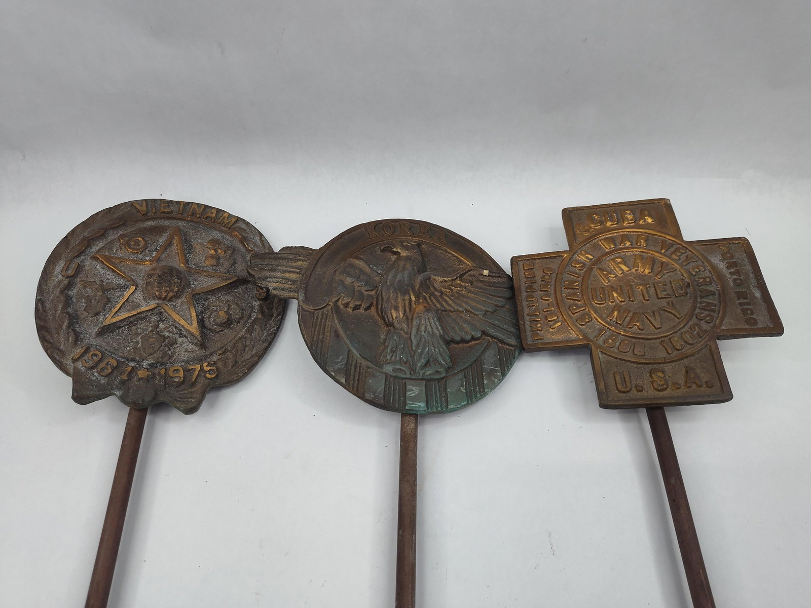 3 American Cast Bronze Grave Markers: This group consists of three cast metal veteran grave markers, likely bronze or brass, each mounted on a tapered metal rod for cemetery placement. The markers correspond to different eras of American