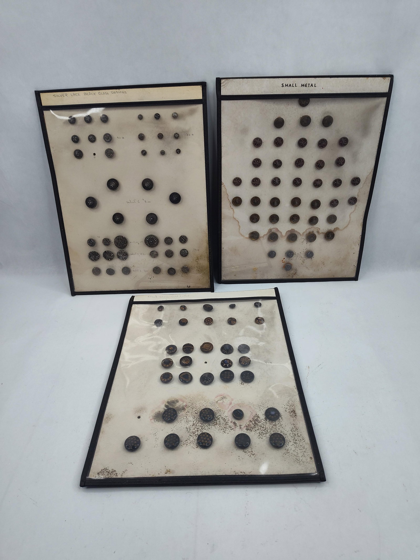 3 Glass & Metal Button Boards: A collection of buttons presented on three individual off-white cardstock display boards housed within black-edged protective covers. The first board features a handwritten header at the top edge