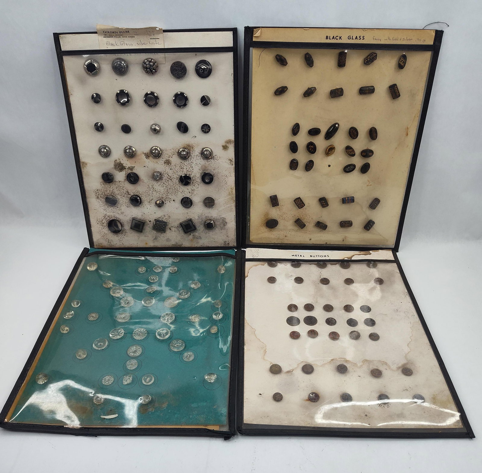 & Glass & Metal Buttons: A collection of buttons presented in four separate black-bordered display folders. The collection is organized by material and finish. One folder contains black glass buttons with a silver luster