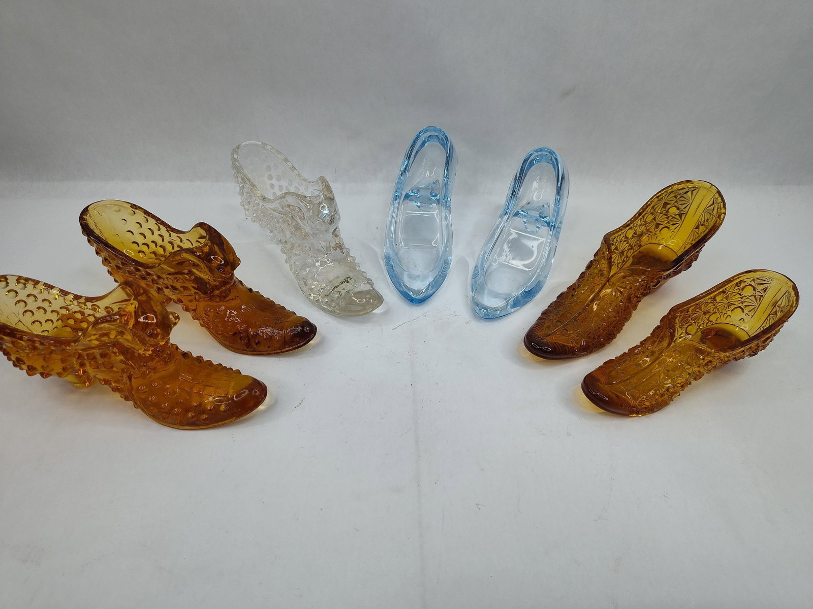 Collection of Pressed Glass Slippers: A collection of decorative pressed glass slippers in various styles and colors. The group includes one clear glass slipper featuring a hobnail pattern and a cat-head motif at the top of the opening.