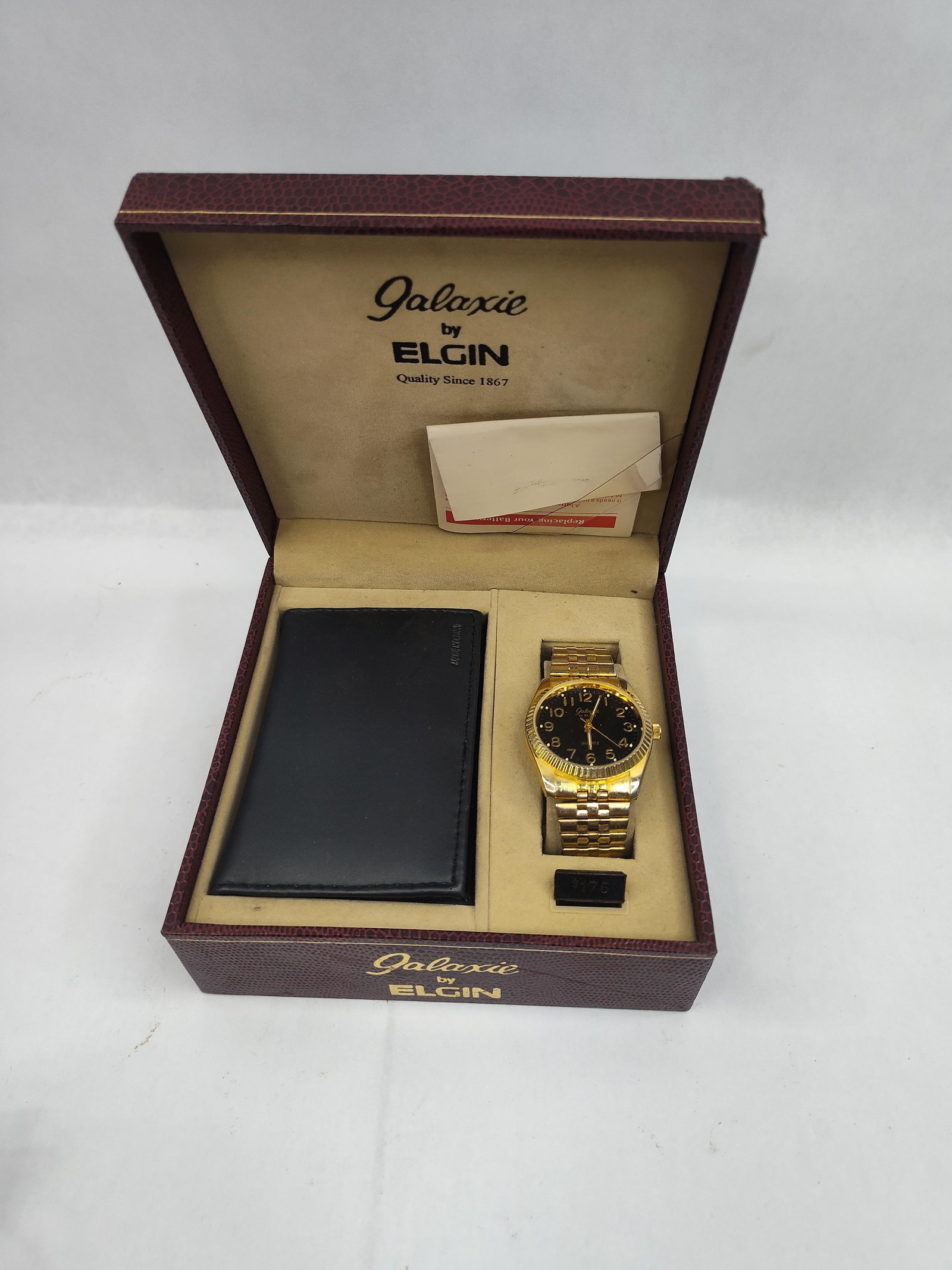 Elgin Galaxie Men's Watch & Wallet Set: A men's gift set presented in a burgundy pebbled-texture hinged display box. The set includes a quartz movement wristwatch and a black bifold wallet. The watch features a gilt-tone case with a fluted 
