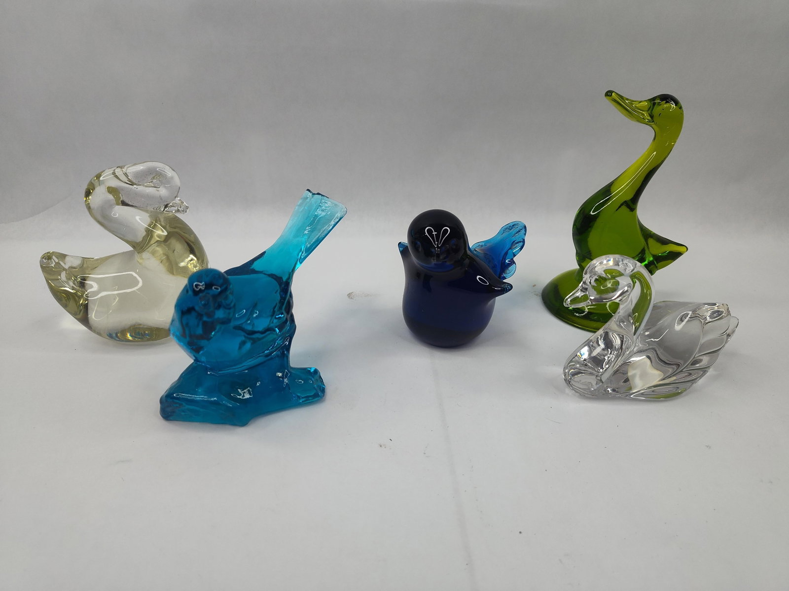 Fenton & Glass Bird Figurine Collection: A collection of glass bird figurines in various colors and styles. The group includes a bright blue bird perched on a textured base, composed of teal-colored glass. An oval white and blue adhesive