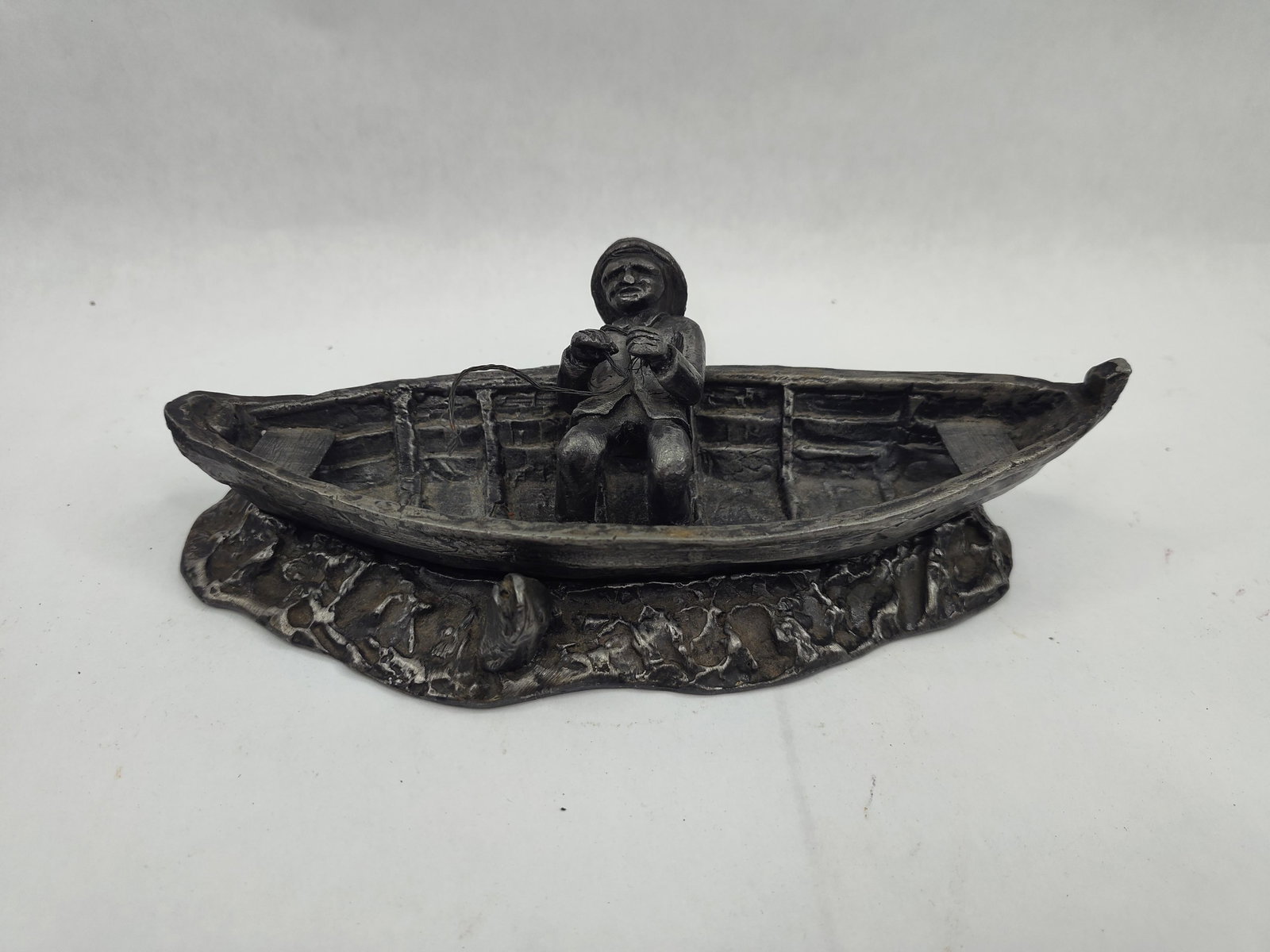Fisherman & Rowboat Metal Sculpture: A three piece cast metal sculptural diorama finished with a dark antiqued patina. The assembly includes a hollow-cast rowboat featuring internal ribbing and two thwarts, a separate seated male figure 