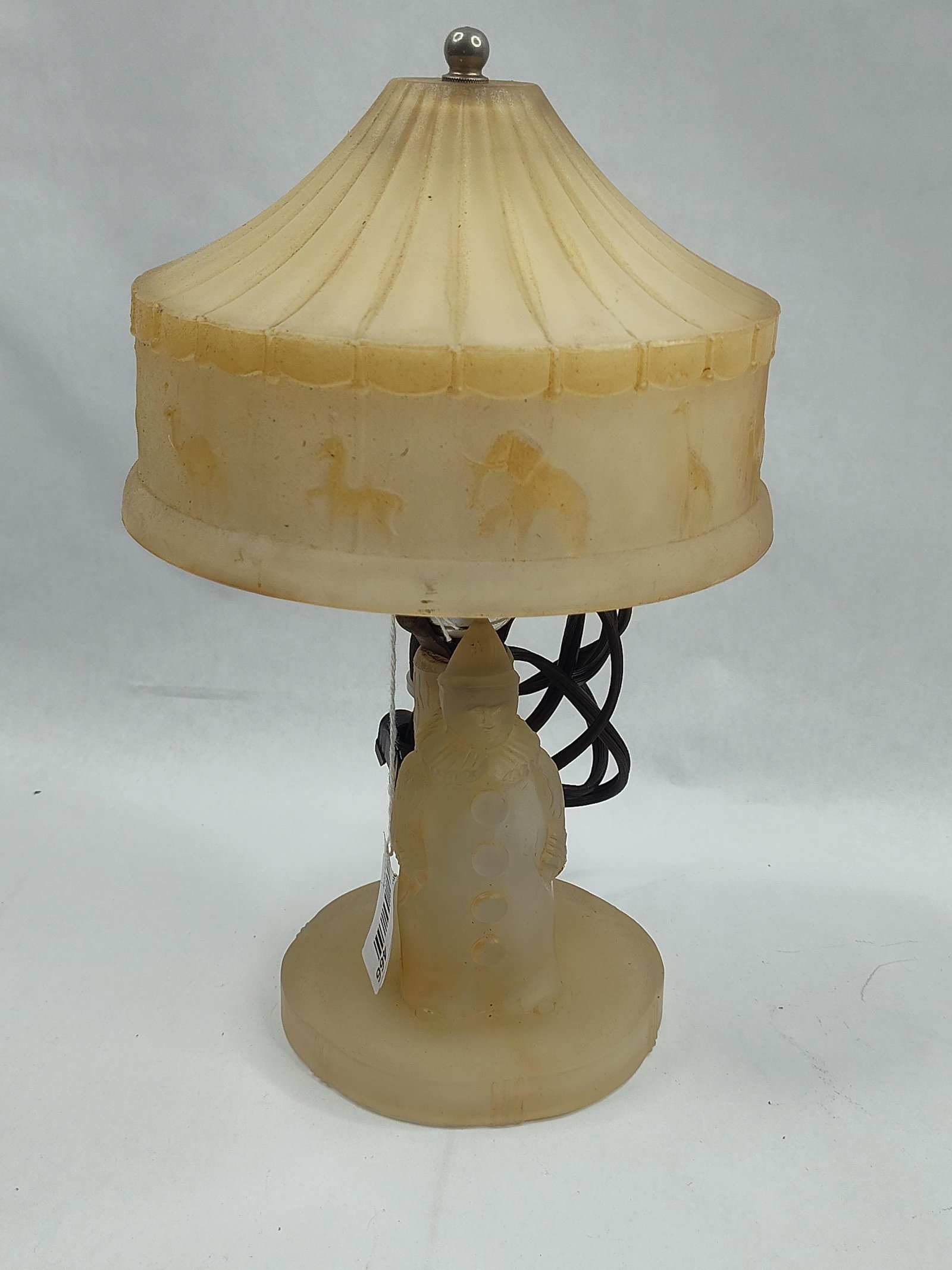 Glass Circus Lamp w/ Clown & Carousel: A mid-20th-century nursery lamp crafted from frosted yellowish-amber glass. The figural base is molded in the form of a standing clown wearing a pointed hat and a ruffled collar, positioned in front