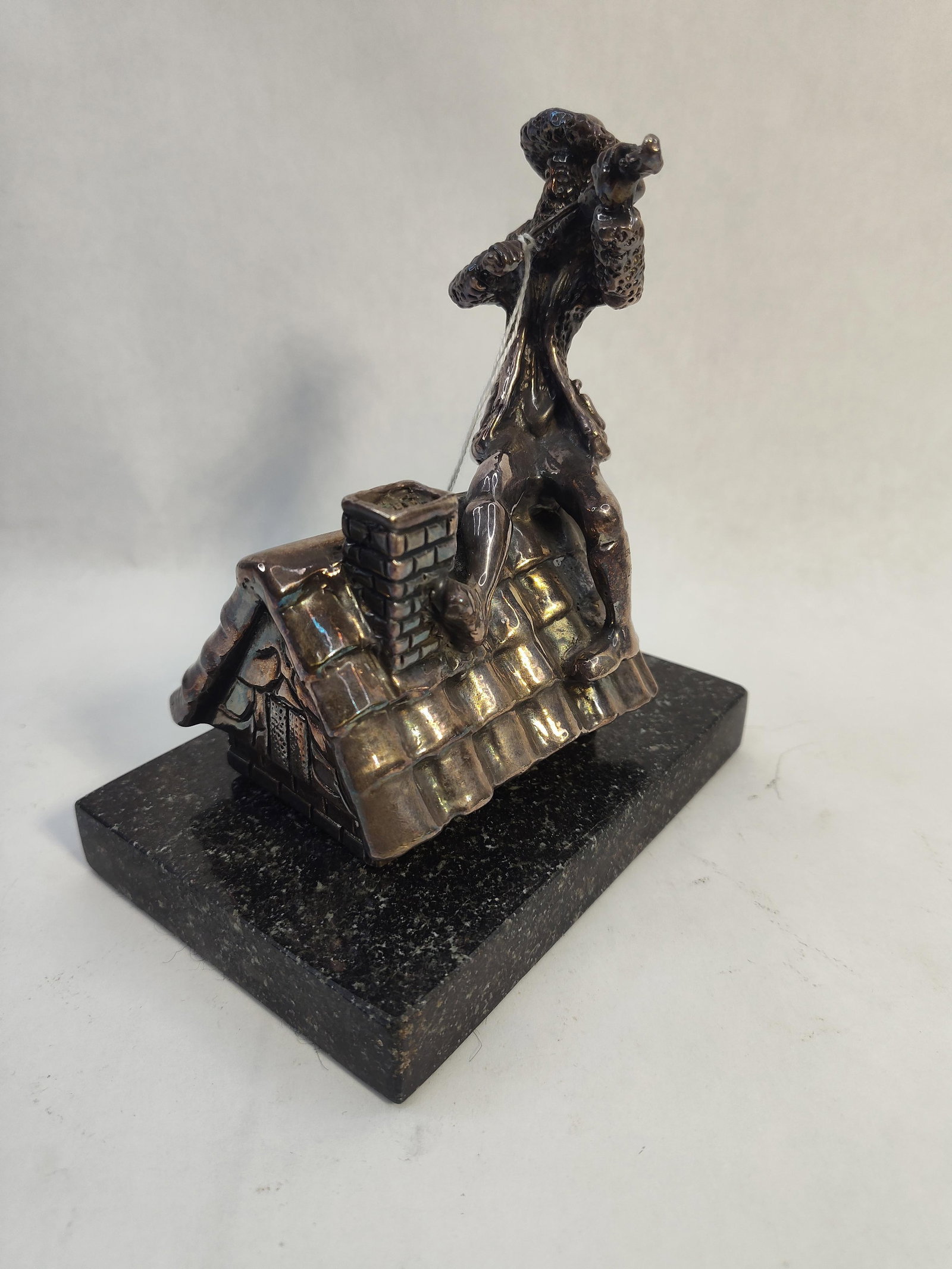 Str. Fiddler on Roof Figurine w/ Marble: This silver figurine depicts the character Tevye from Fiddler on the Roof, characterized by his beard, traditional cap, and long coat. He is shown in a seated position atop a pitched roof with a chimn