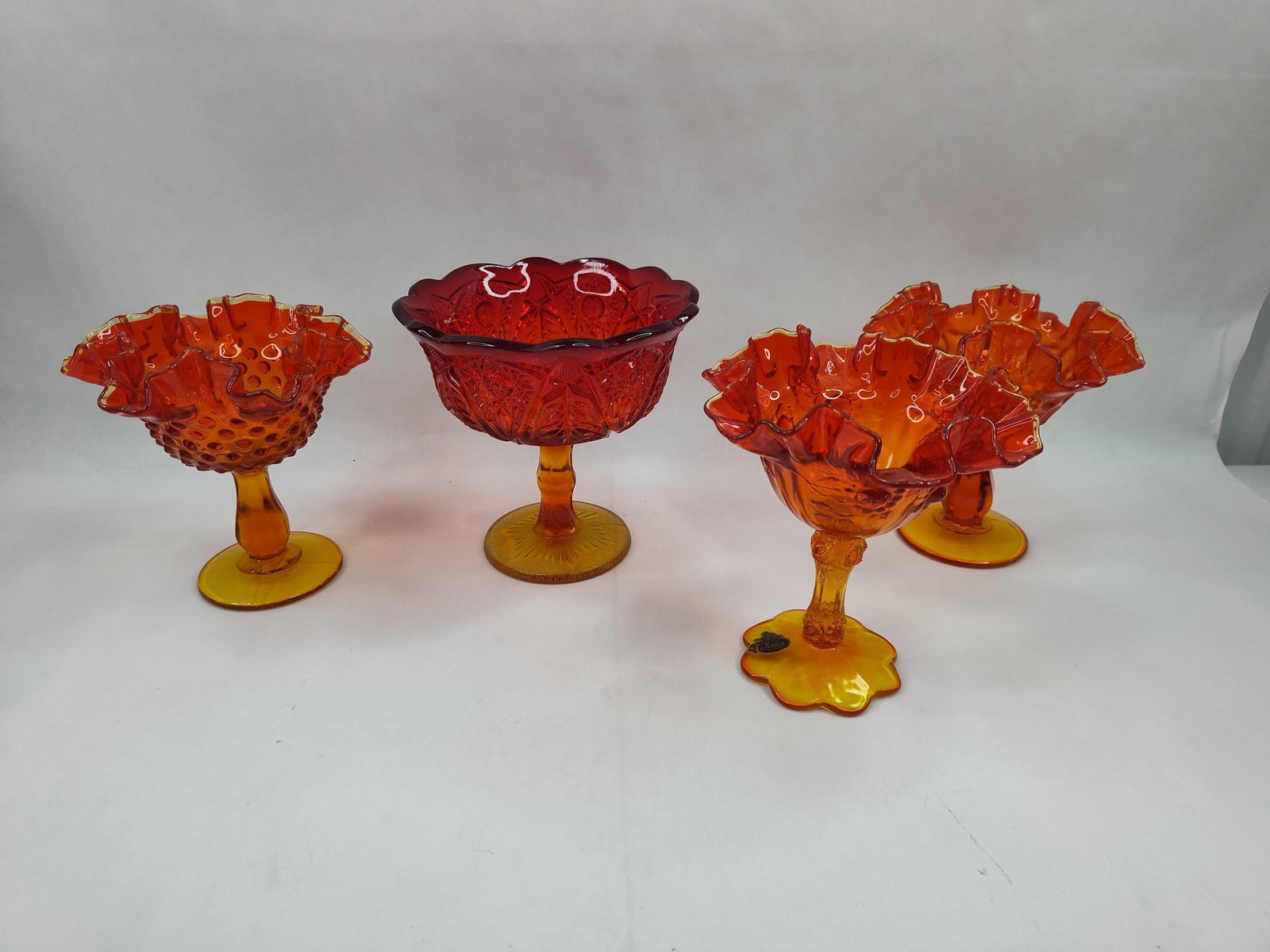 Fenton Amberina Glass Bowls & Compotes: A collection of American art glass pieces featuring Amberina coloration, defined by a fire-orange and yellow-amber gradient transitioning to deep ruby red at the rims. The group includes several