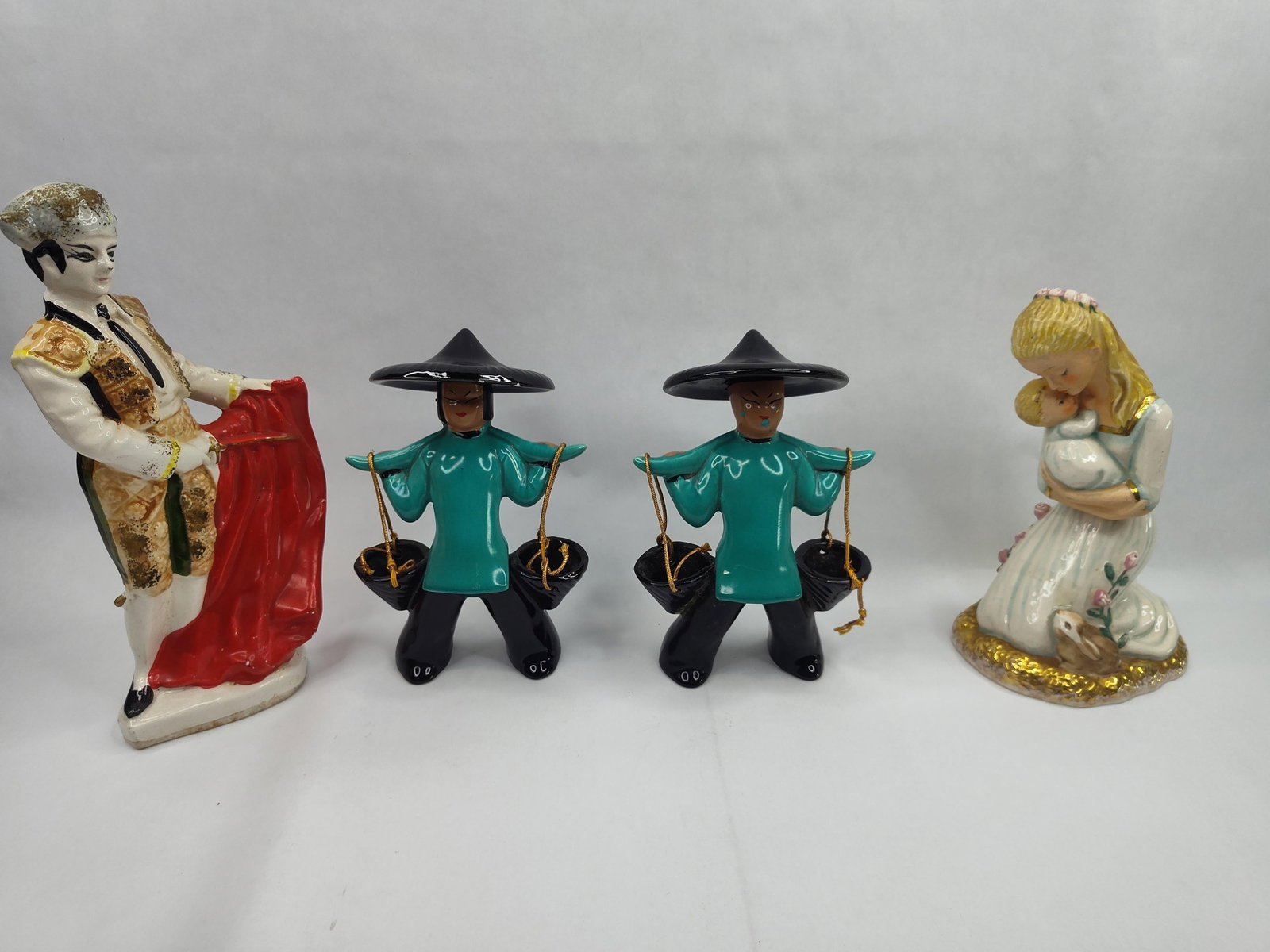 Goebel & Glazed Ceramic Figurine Set: A collection of four glazed ceramic figurines of varying styles. The collection includes a West German kneeling mother and child figure, a pair of turquoise-clad water carriers, and a Spanish