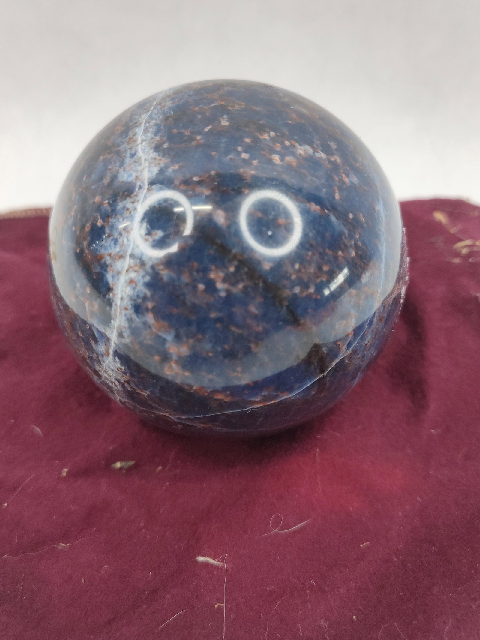 Blue Sodalite Lapidary Sphere: A polished lapidary sphere composed of sodalite, characterized by a deep navy blue matrix. The stone exhibits natural inclusions of reddish-brown feldspar and white quartz, appearing as a mottled and