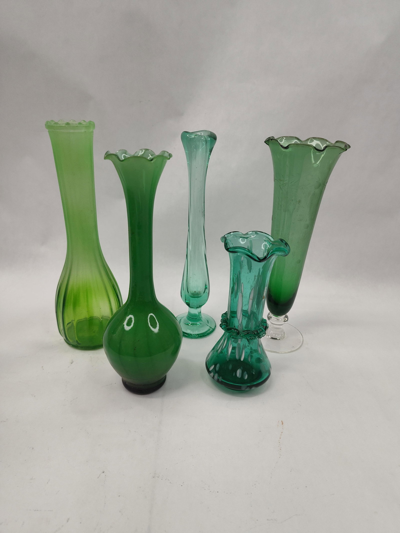 An Assortment of Green Glass Bud Vases: A collection of various green glass bud vases representing a range of mid-century glassmaking styles and techniques. The group includes a tall light green vase with a translucent satin finish,