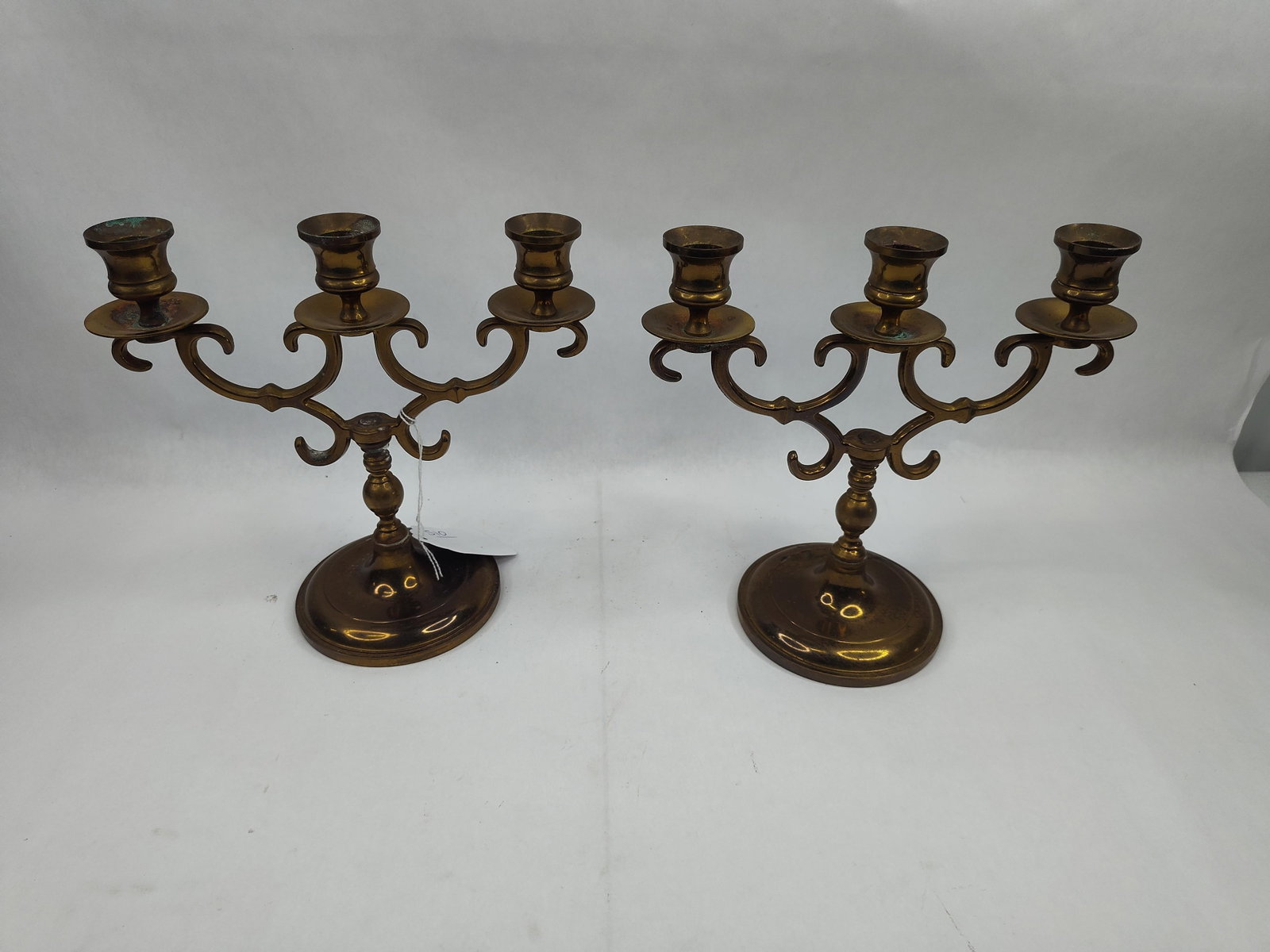 Pair of 3-Light Brass Candelabra: A pair of three-light brass candelabra constructed with central shafts featuring baluster turnings. Each candelabrum supports two scrolling, openwork arms that terminate in circular drip pans and