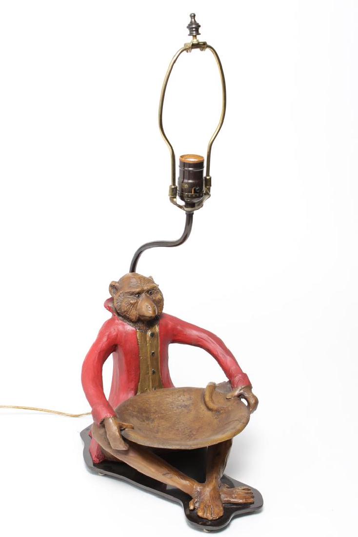 Bill Huebbe-Manner Cast Monkey Table Lamp: Cast table lamp of a seated monkey in red frock coat and gilt vest holding a bowl, in the style of Bill Huebbe, mounted on a black metal base. 24.5" H. Small chip to back of coat collar.