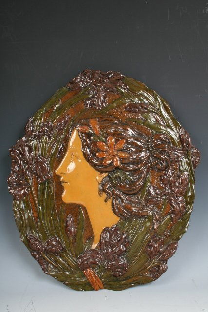 Wahliss Nouveau Ceramic Plaque Woman w/Lilies: Art-Nouveau-style polychrome ceramic oval plaque showing the profile head of a young woman, modeled in high relief within a vignette of lilies, c. 1900. Marked Ernst Wahliss on the verso with a serial