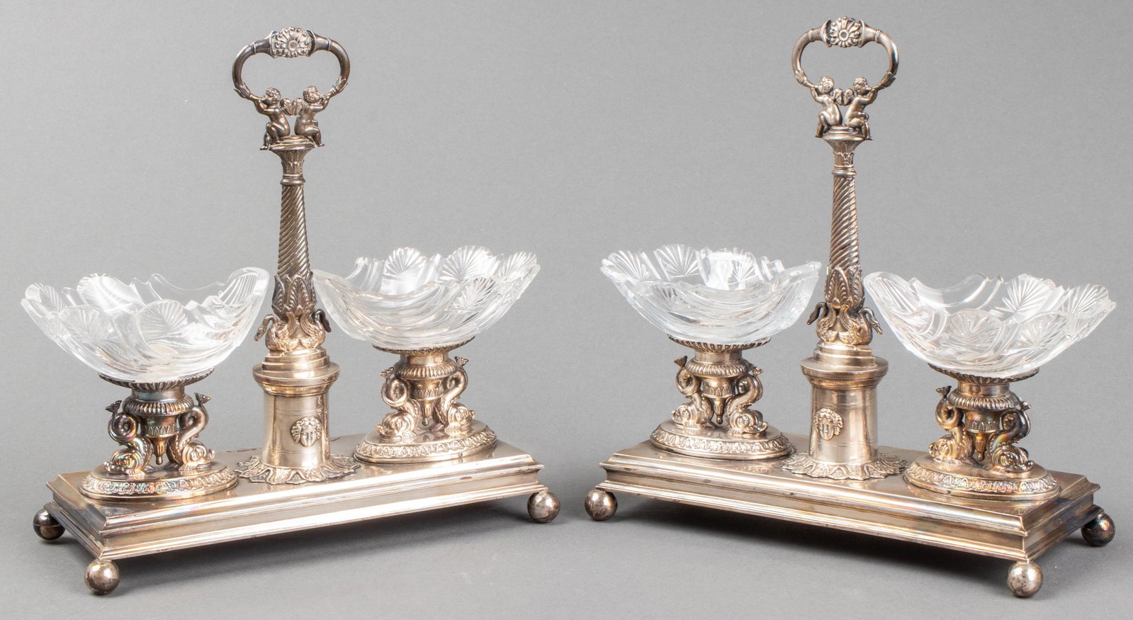Rococo Style Candy Dish Stands, Pair: Pair of Rococo style silver-plated stands with glass candy or nut dishes, throughout with dolphin, cherub, swan, and acanthus motifs, raised on ball feet, unmarked. 7.5” H x 8” W x 3” D. Provena