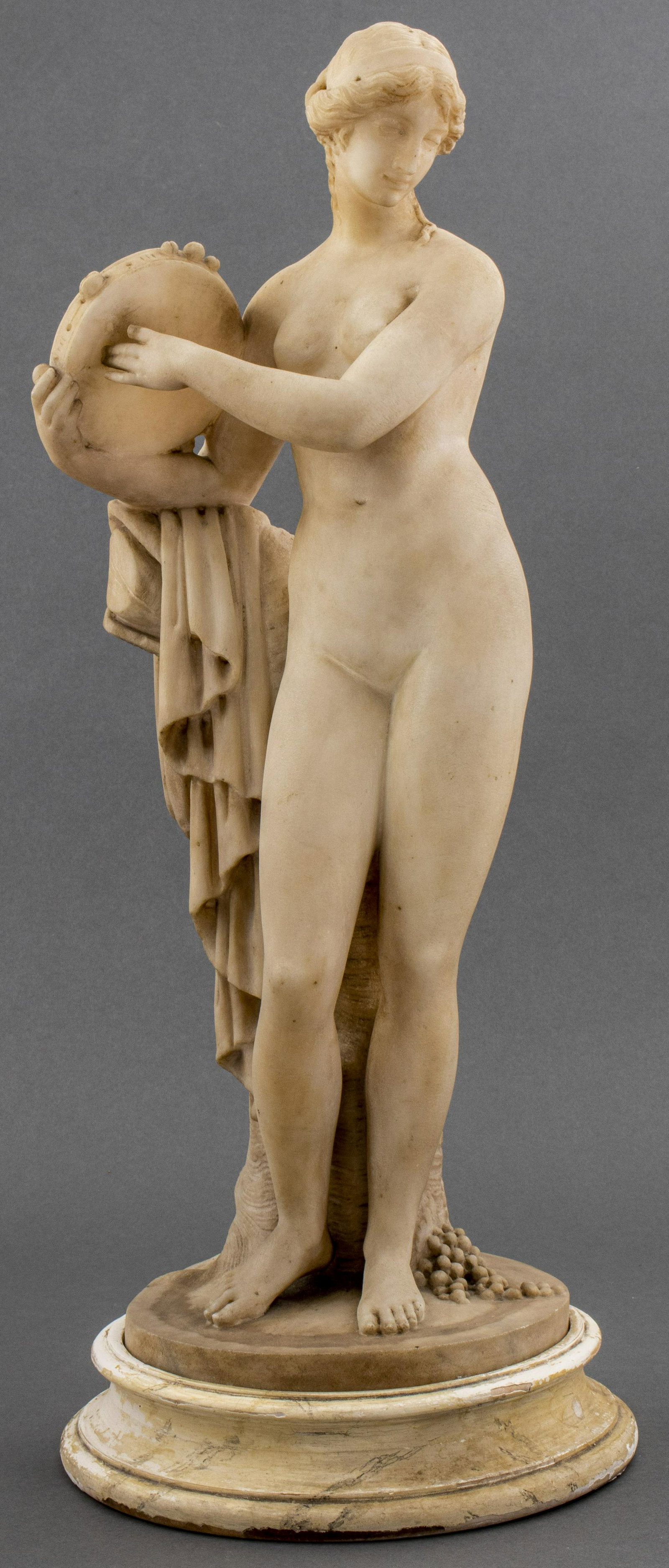Classical Style Carved Alabaster Statue: Classical style carved alabaster statue, after the antique, modeled as a nude maiden with a tambourine against a tree trunk, on a round base. Overall 22.25” H x 9” diameter. Provenance: Pr