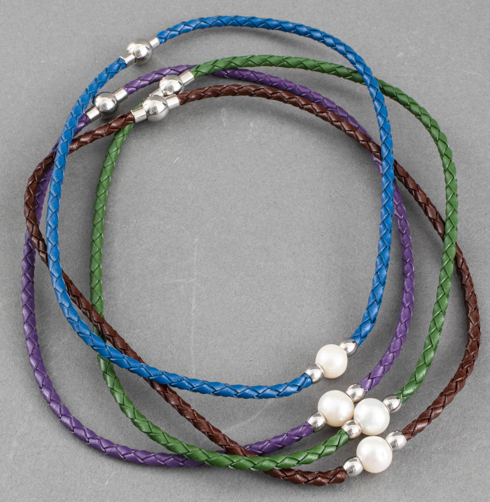 Contemporary Single Oval Pearl Leather Chokers: Four contemporary braided leather cord chokers with silver-tone fittings bearing a single ringed oval pearl and magnetic clasps. All unmarked. Cord colors blue, brown, purple, and green. Pearls approx
