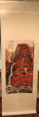 Chinese Landscape Scroll Painting by Li Keran: Scroll painting picturing clusters of houses on a cliffside of a red, mountainous landscape above a rocky river fed by a waterfall on the left side of the painting, by Li Keran (1909-1989), ink on pap