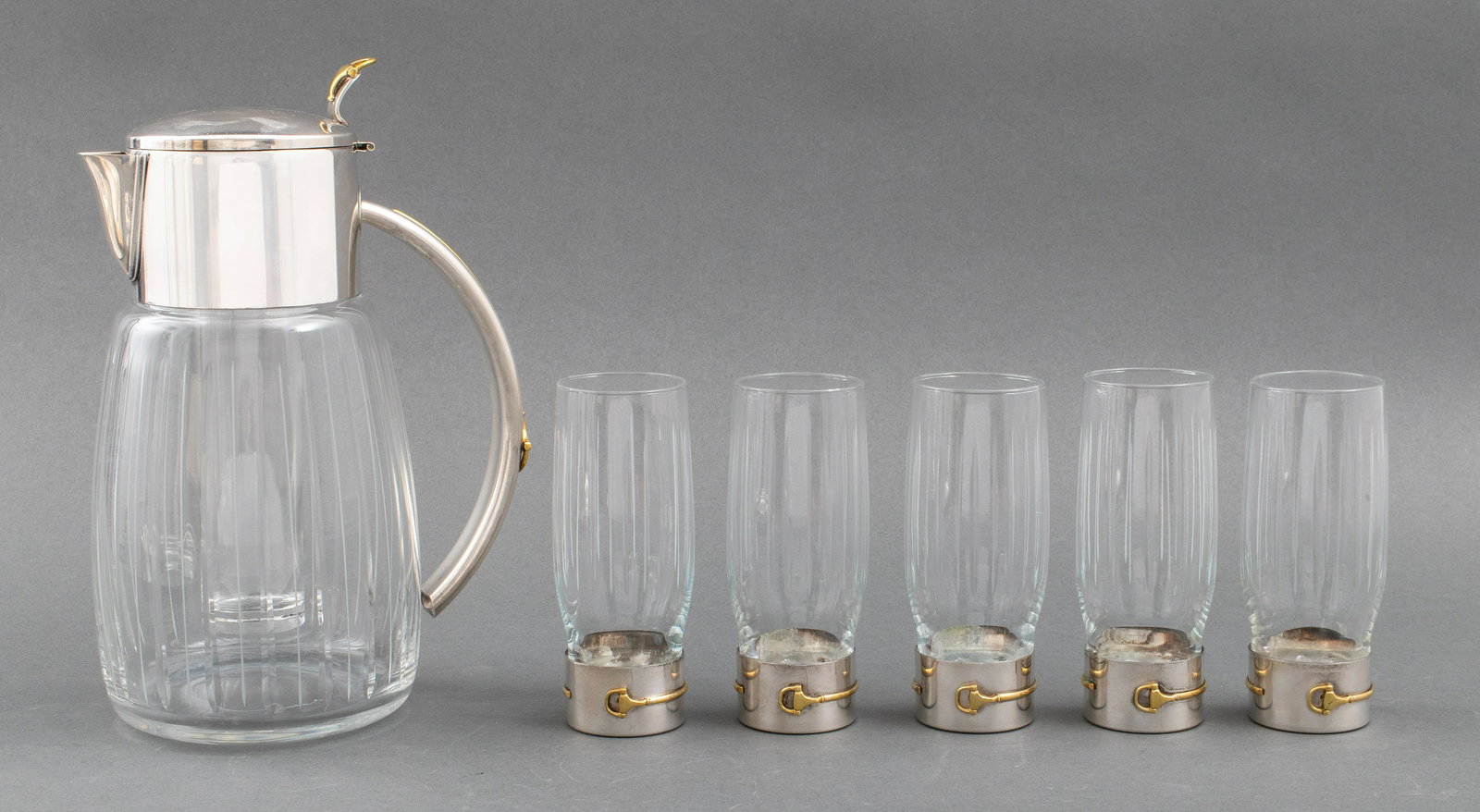 Gucci Stainless Steel, Brass & Glass Drink Service: Gucci modern stainless steel, brass & glass drinks service, comprising five brass and steel mounted glasses, and a stainless steel and glass mounted thermal pitcher, the bottom of the glasses struck "