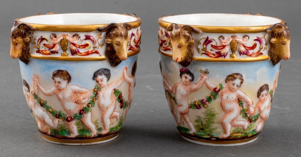 Capodimonte Tete de Belier Cups, 2: Pair of Neapolitan Capodimonte beakers or cups, each with underglaze blue crowned "N" mark, and each with "tete de belier" or ram's head ornamentation to rim, and polychromed cherub decoration to side
