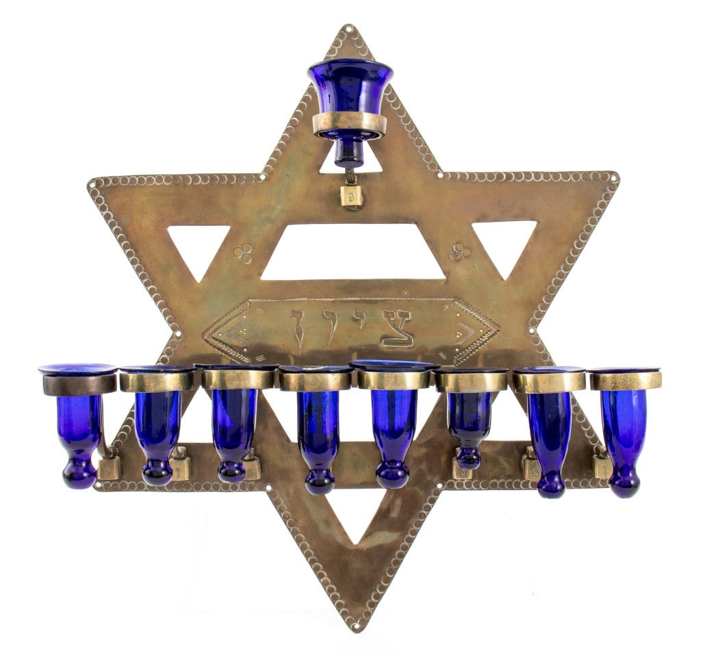 Judaica Brass Star of David Menorah: Judaica Brass Star of David Menorah, incised with Hebrew, with nine cobalt blue glass oil cups. One glass cup with minor loss to rim. 18.25" H x 16.5" W x 5" D. Provenance: From a 25 E. 83rd Street es