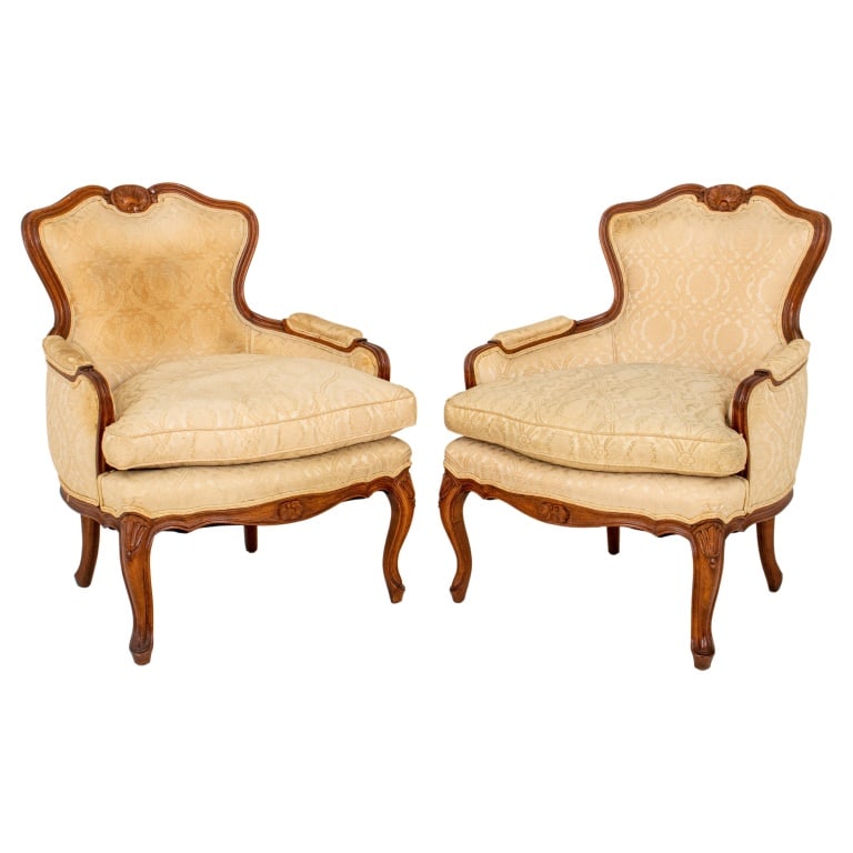 Louis XV Style Upholstered Bergeres, Pair: Pair of Louis XV Style Bergeres, carved fruitwood with shell patterns on back, and a shaped seat with cushion, upholstered in a beige damask fabric, above cabriole legs with scroll feet. 34.5" H x 26.